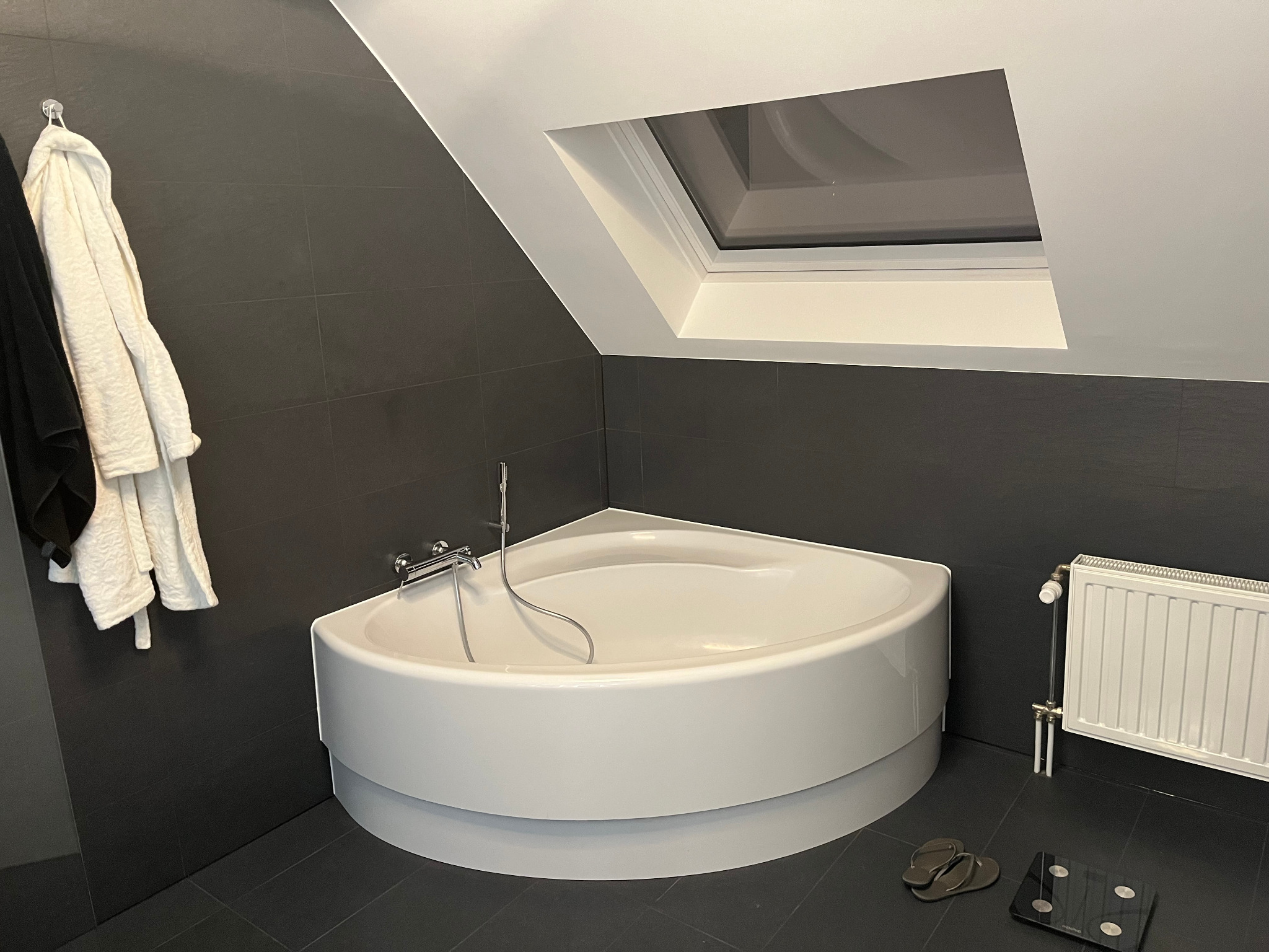 Nowoczesna łazienka na poddaszu z narożną wanną, czarnymi płytkami i oknem dachowym. Minimalistyczny design z białym grzejnikiem i ręcznikami.