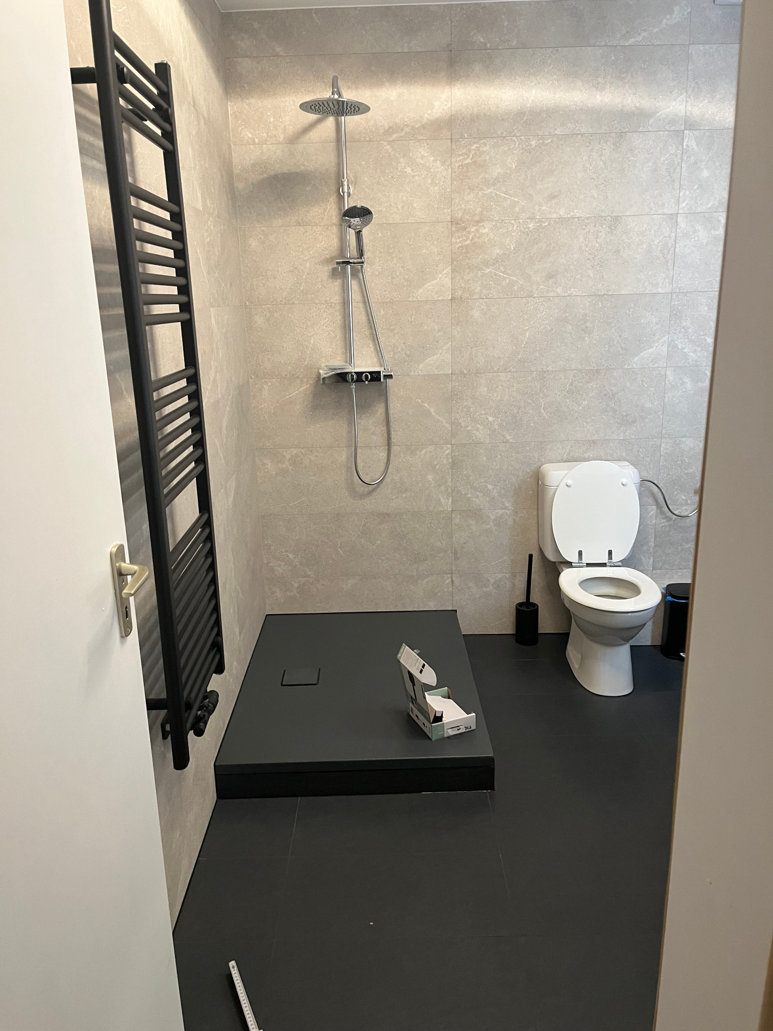 Wykończona łazienka z czarnym brodzikiem, grzejnikiem drabinkowym i beżowymi płytkami. Widoczna toaleta i akcesoria łazienkowe. Częściowo widoczna miarka na podłodze.