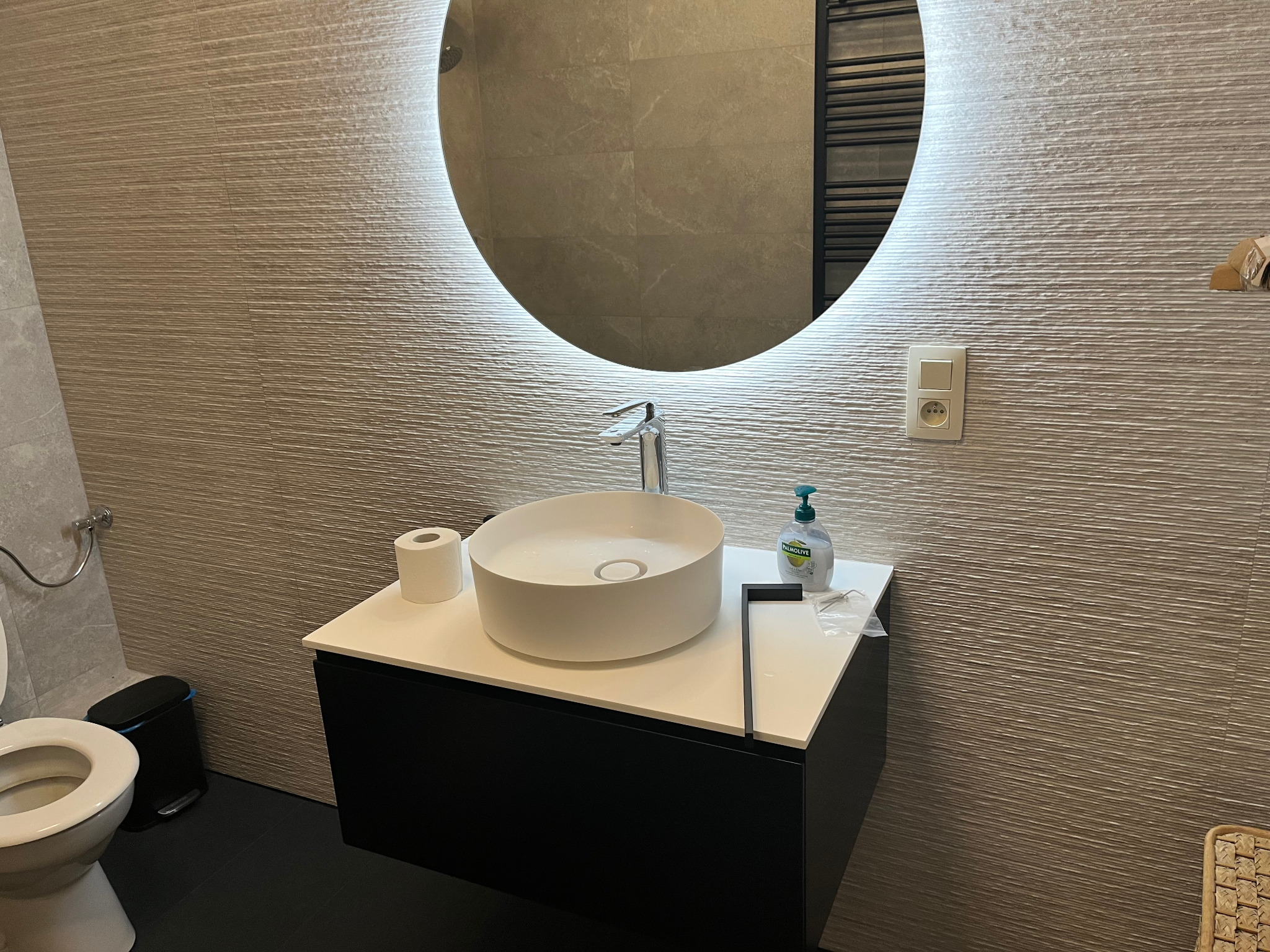 Nowoczesna łazienka z okrągłym lustrem podświetlanym LED, umywalką nablatową i teksturowaną ścianą. Minimalistyczny design, czarne akcenty i subtelne oświetlenie.