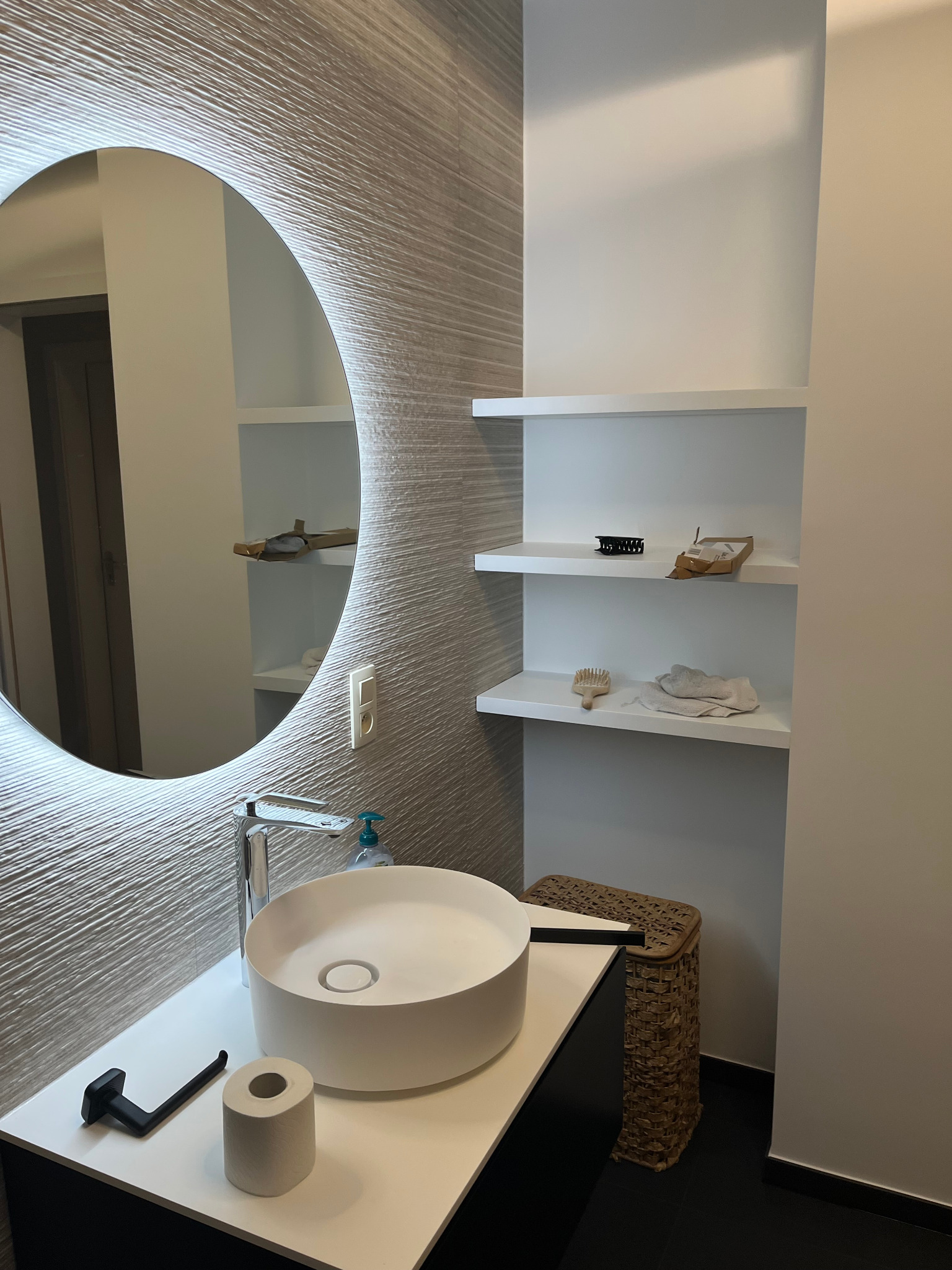 Nowoczesna łazienka z okrągłym lustrem z podświetleniem LED, umywalką nablatową i półkami wnękowymi. Minimalistyczny design, beżowo-biała kolorystyka.