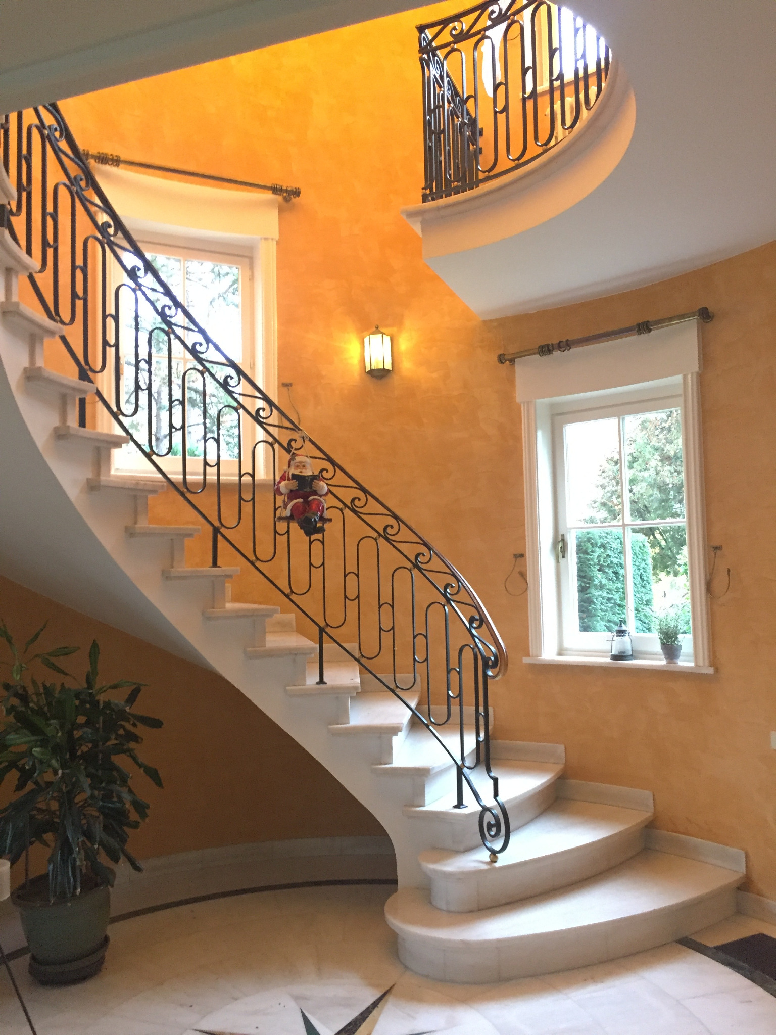 Eleganckie schody spiralne z kutą balustradą i marmurowymi stopniami, oświetlone ciepłym światłem, na tle ściany w odcieniach pomarańczy. Dekoracja świąteczna na balustradzie.