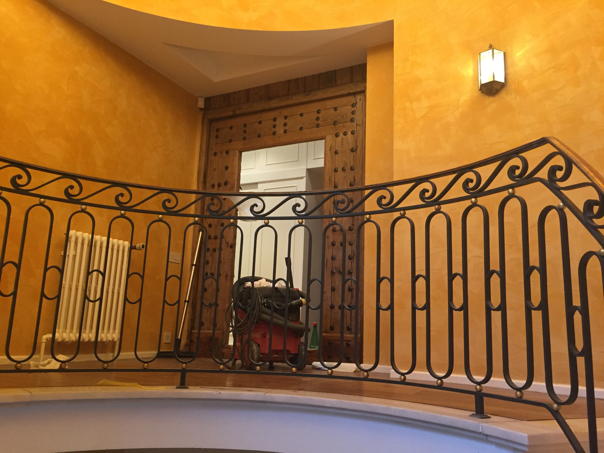 Klatka schodowa z dekoracyjną balustradą i ścianami w odcieniach złota. Widoczny grzejnik, drewniane drzwi z metalowymi zdobieniami i lampa ścienna.