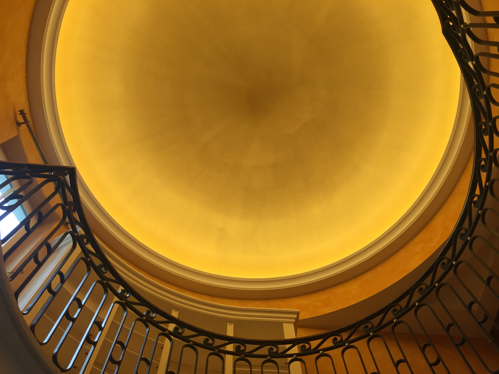 Złota kopuła sufitu z oświetleniem w niszach, widoczna z dołu przez ozdobną, metalową balustradę schodów. Ciepłe światło podkreśla fakturę ścian.