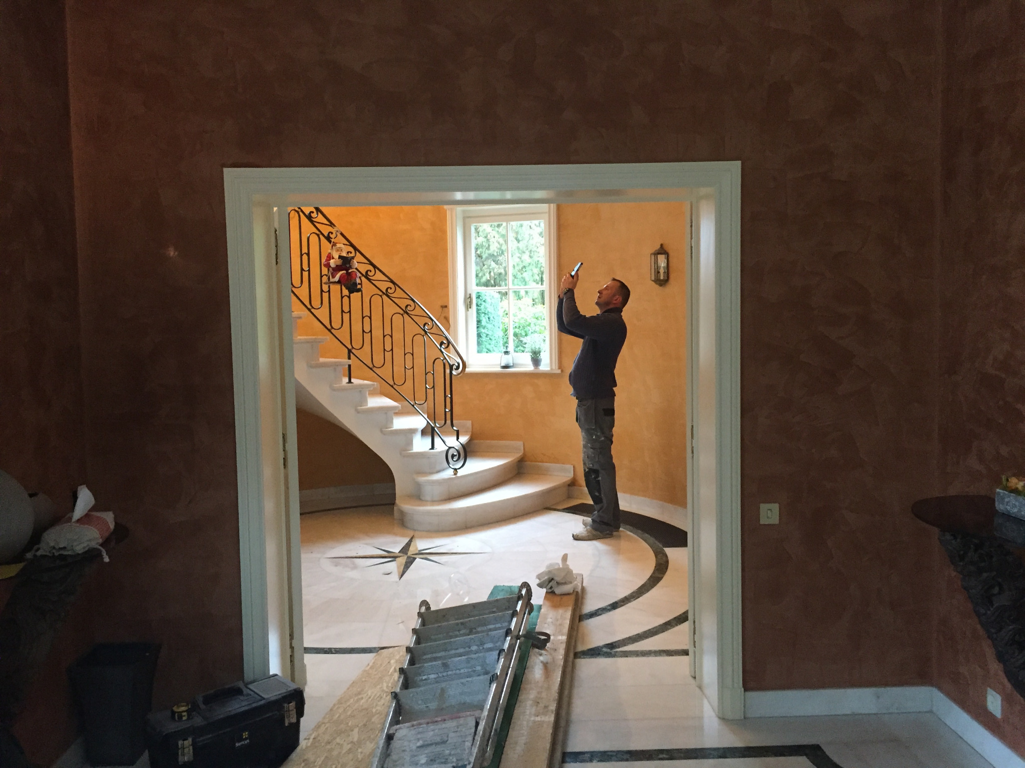 Wnętrze z marmurową podłogą i schodami, widoczne przez drzwi. Mężczyzna robi zdjęcie smartfonem. Ściany w odcieniach brązu i pomarańczy, dekoracyjna sztukateria.
