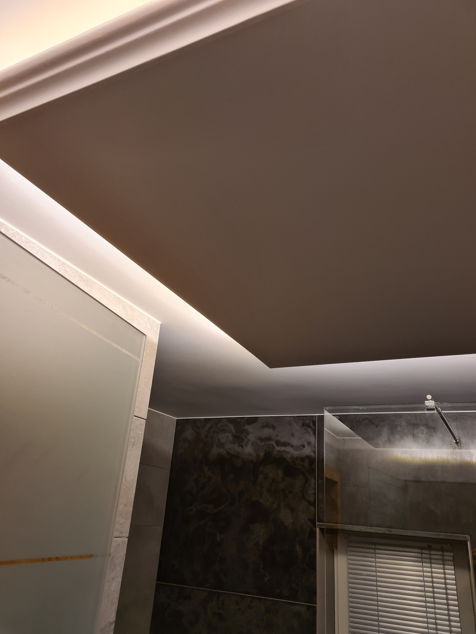 Nowoczesna łazienka z podwieszanym sufitem i oświetleniem LED. Widoczne szklane elementy kabiny prysznicowej i ciemne płytki imitujące marmur.