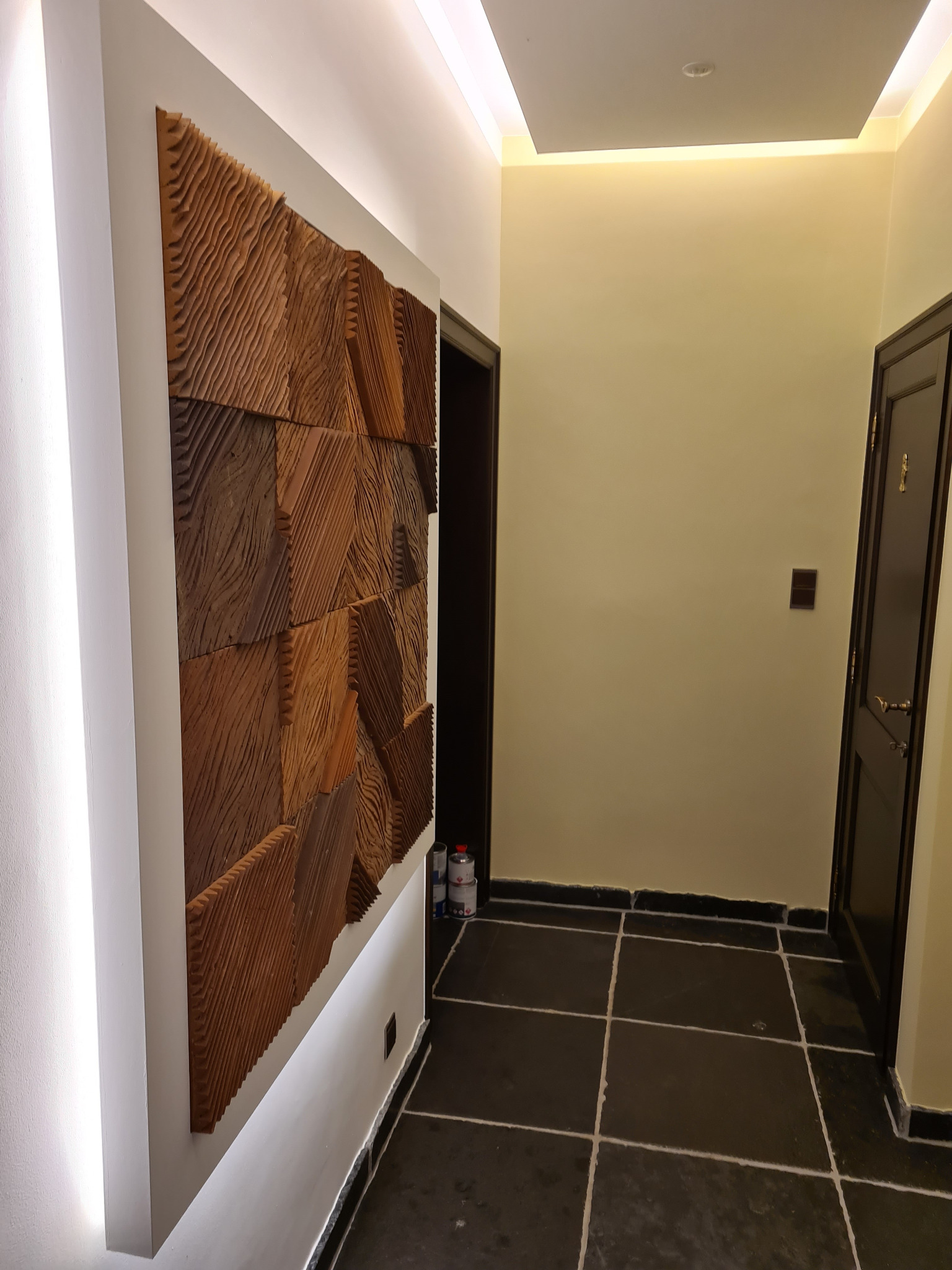 Korytarz z dekoracyjną, drewnianą płaskorzeźbą na ścianie, podświetloną listwą LED. Ciemna podłoga z płytek, drzwi po prawej stronie. Nowoczesne wykończenie.