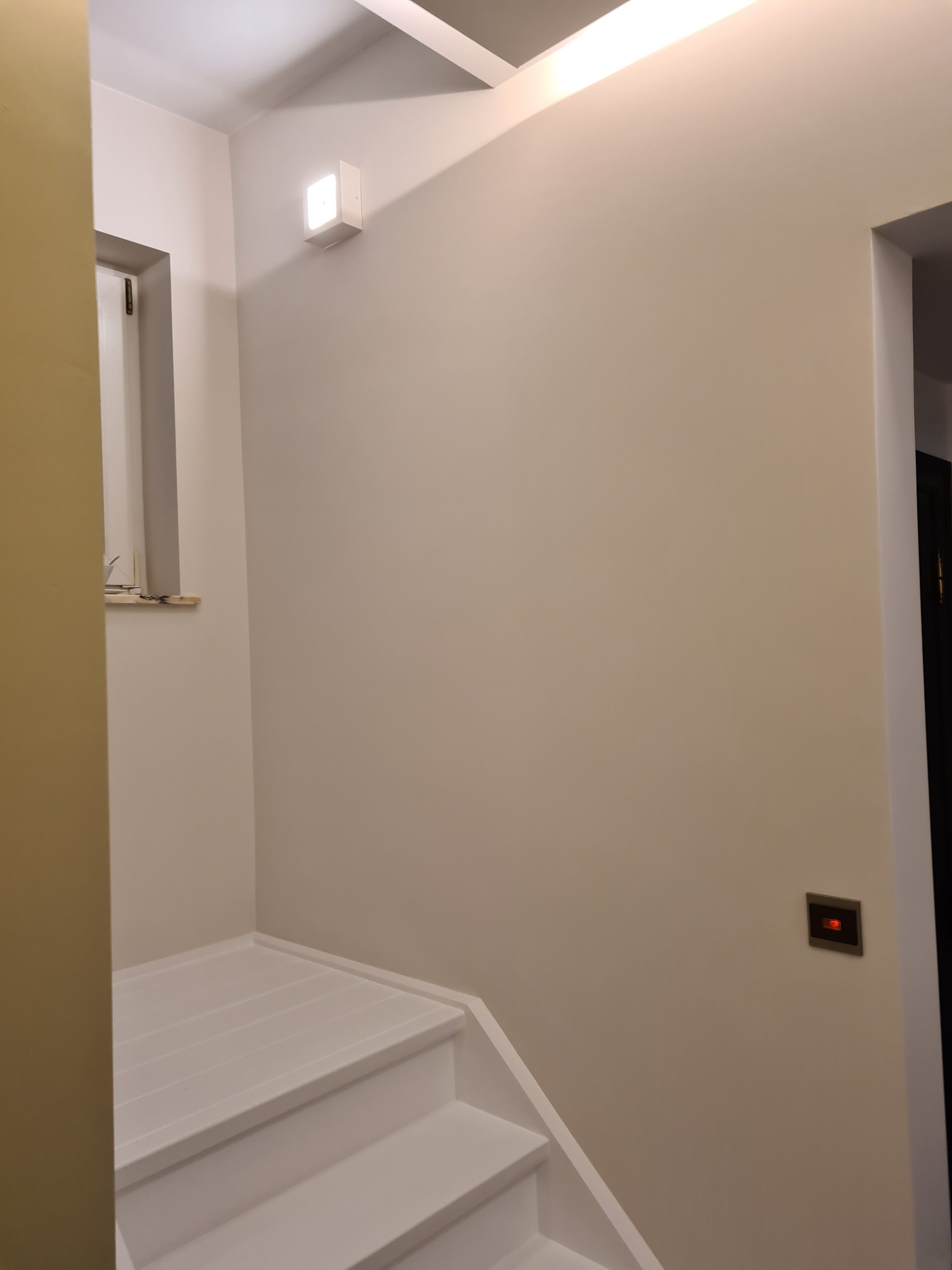 Jasne wnętrze po odnowieniu: białe schody, kremowe ściany, nowoczesne oświetlenie i minimalistyczny włącznik. Widok z dołu, podkreślający świeżość i prostotę.