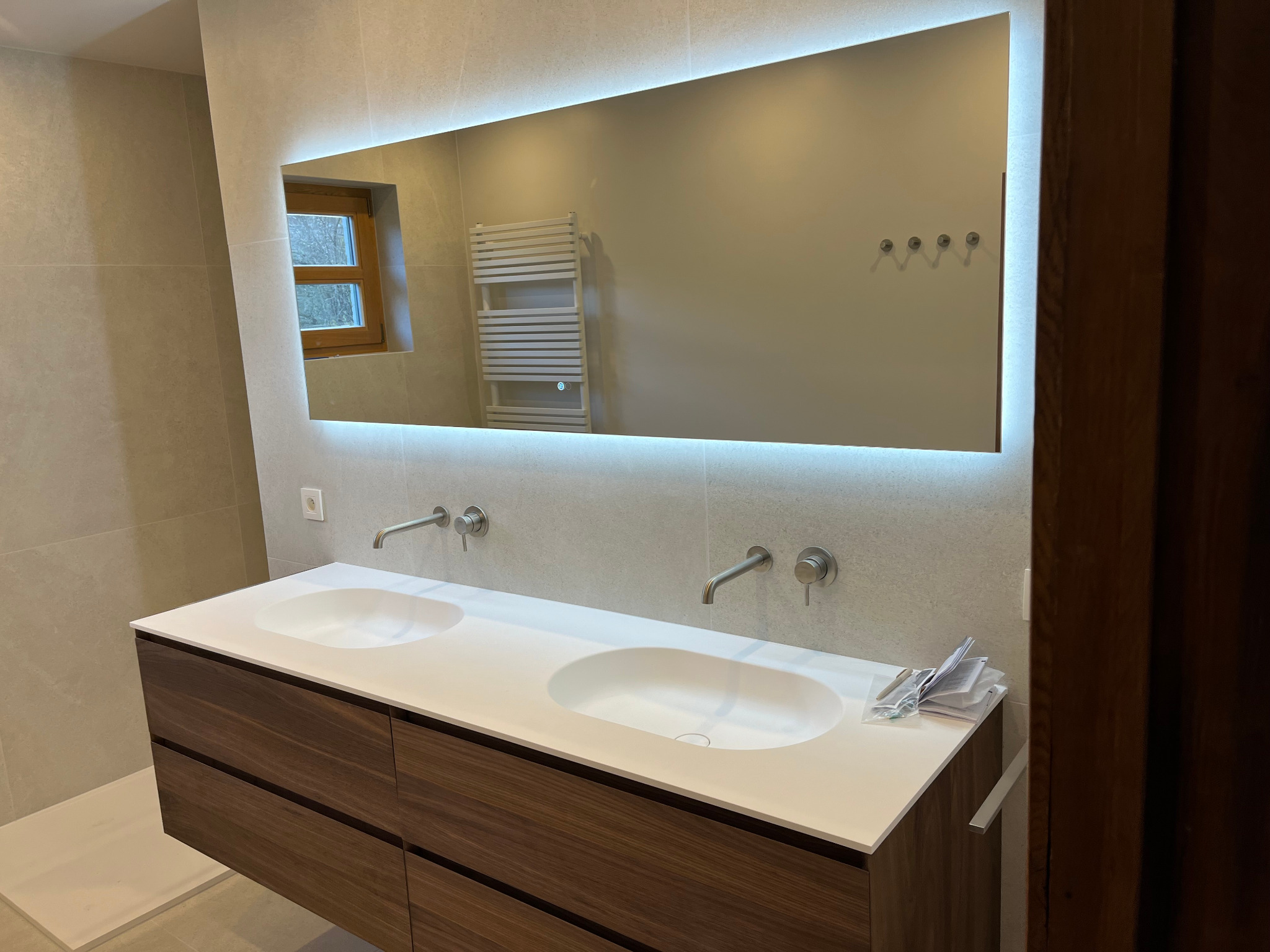 Nowoczesna łazienka z podwójną umywalką na drewnianej szafce, duże lustro z podświetleniem LED i beżowe płytki na ścianach. Widoczne baterie i akcesoria.