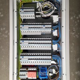 El-Home Piotr Janucik - Rozdzielnica elektryczna w białej obudowie, z widocznymi bezpiecznikami, przewodami i elementami sterującymi. Instalacja schludna, z dbałością o detale.