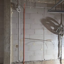 El-Home Piotr Janucik - Surowa ściana z betonu komórkowego z poprowadzonymi przewodami elektrycznymi w peszlach i kablami. Widoczne puszki instalacyjne i fragmenty konstrukcji sufitu.