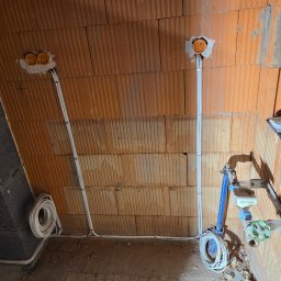 El-Home Piotr Janucik - Surowa ściana z cegły z widocznymi puszkami instalacyjnymi i przewodami elektrycznymi. Instalacja w trakcie montażu, widoczne elementy instalacji wodnej.
