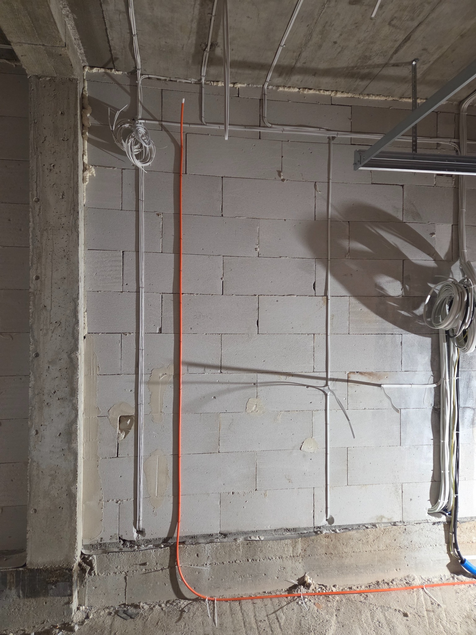 Surowa ściana z betonu komórkowego z poprowadzonymi przewodami elektrycznymi w peszlach i kablami. Widoczne puszki instalacyjne i fragmenty konstrukcji sufitu.