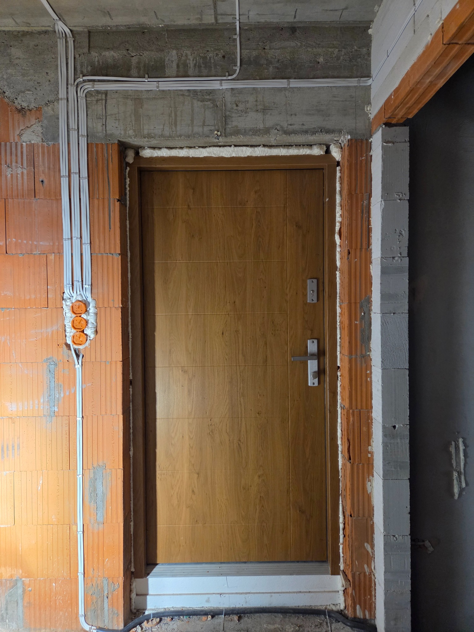 Montaż drzwi wejściowych w surowym stanie, widoczne przewody elektryczne i puszki instalacyjne w ścianie z cegły. Pianka montażowa wokół ościeżnicy.