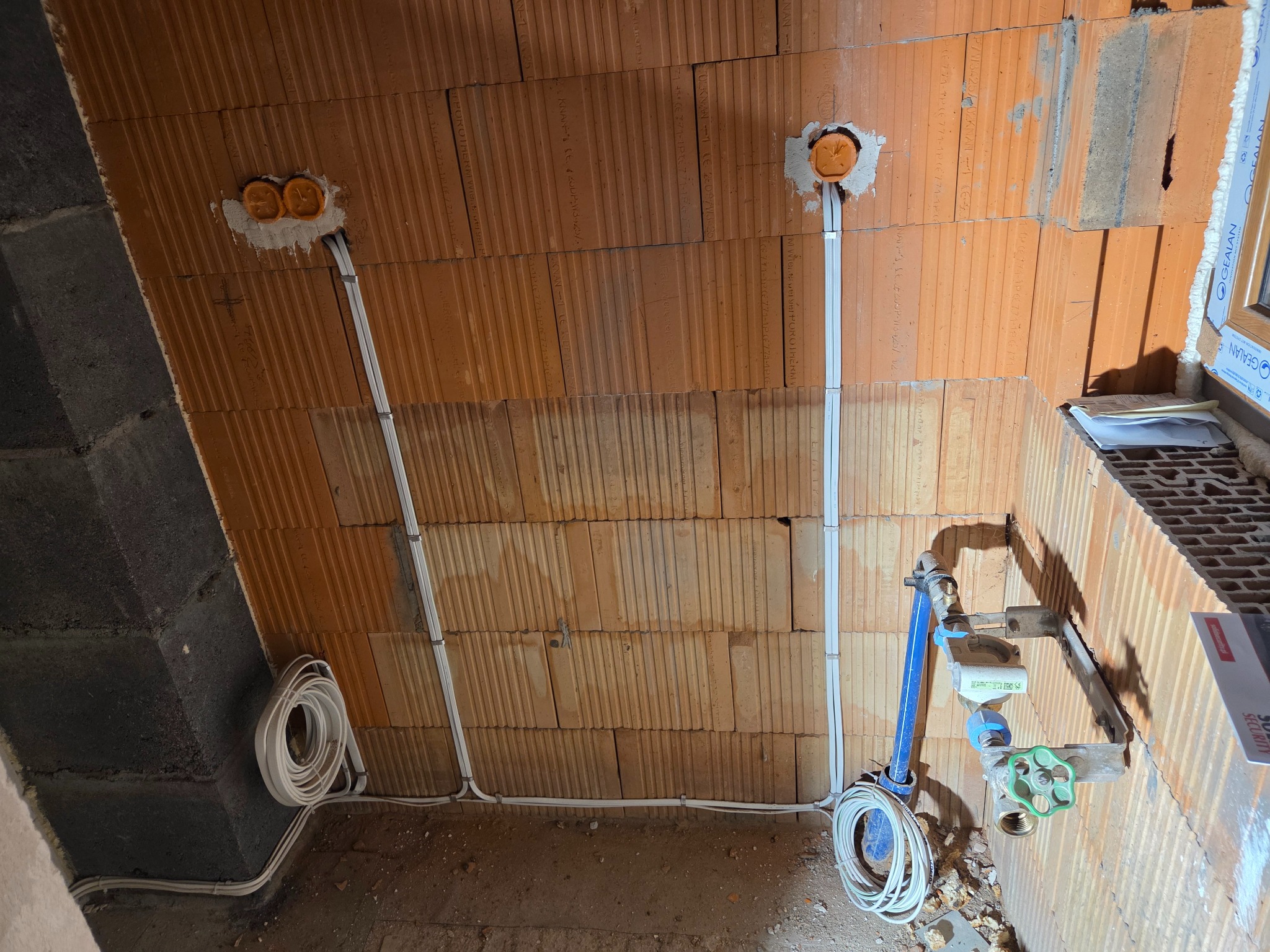 Surowa ściana z cegły z widocznymi puszkami instalacyjnymi i przewodami elektrycznymi. Instalacja w trakcie montażu, widoczne elementy instalacji wodnej.