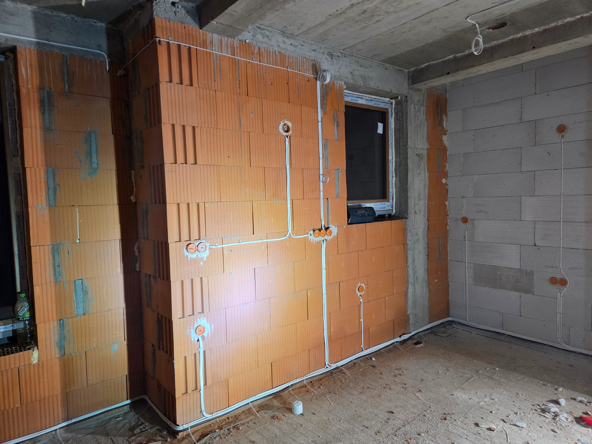 Wnętrze w trakcie prac: pomarańczowe cegły z widoczną instalacją elektryczną w białych rurkach, gniazdka w puszkach, surowy betonowy strop, okno z butelką wody.