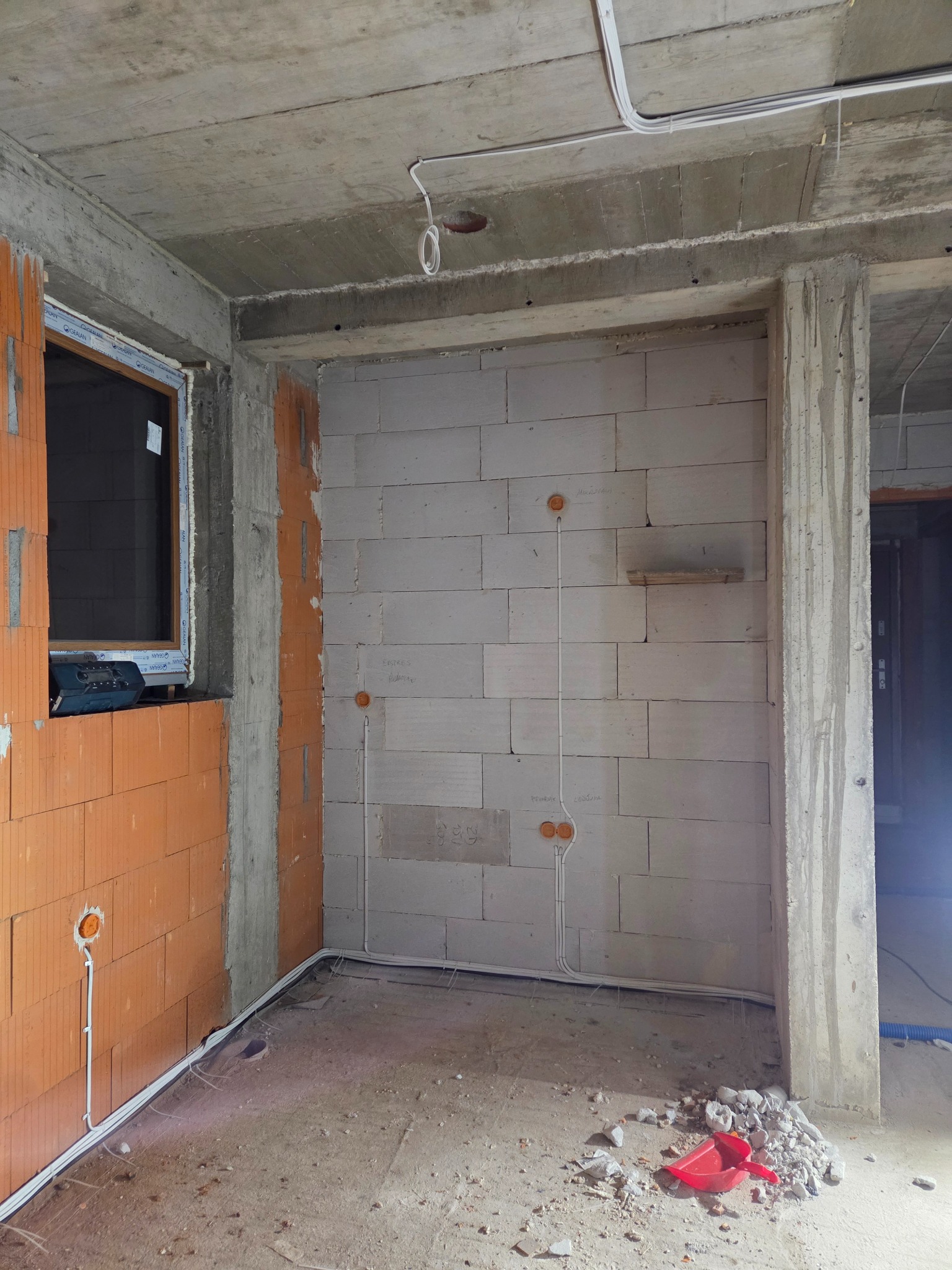 Wnętrze w trakcie prac elektrycznych: widoczne przewody w ścianach z pustaków, puszki instalacyjne, okno z narzędziem, surowy betonowy strop i resztki gruzu na podłodze.