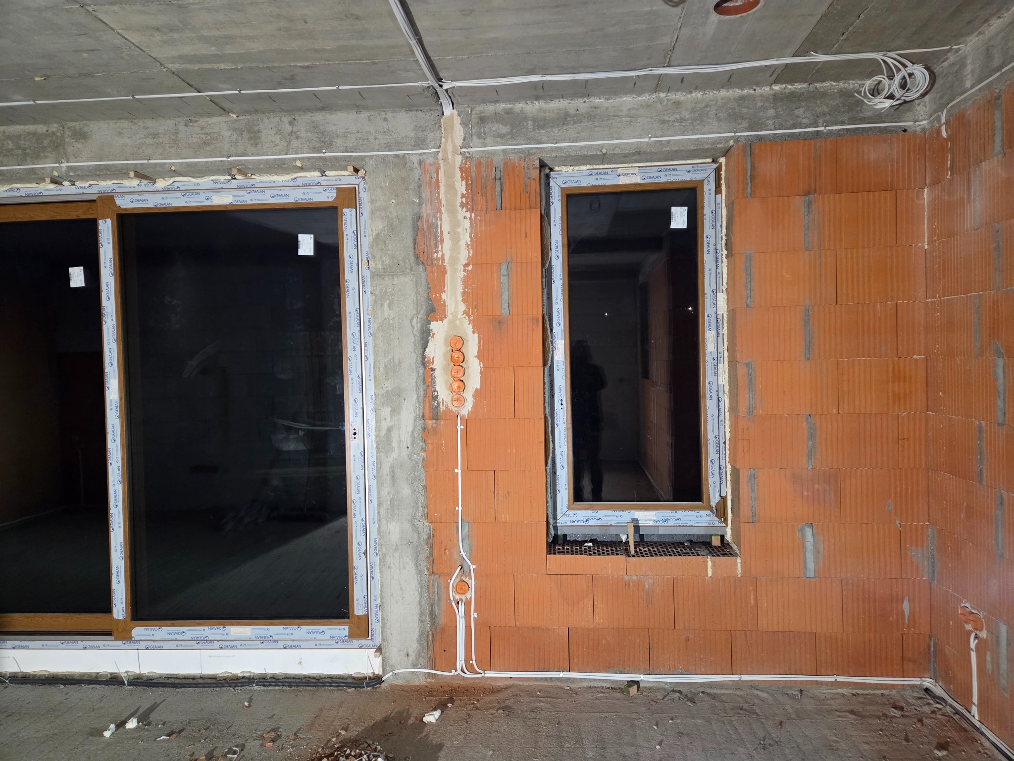 Surowy stan: Instalacja elektryczna w nowym budynku z widocznymi przewodami i puszkami podtynkowymi na tle ściany z cegły i okien z profili Gealan.