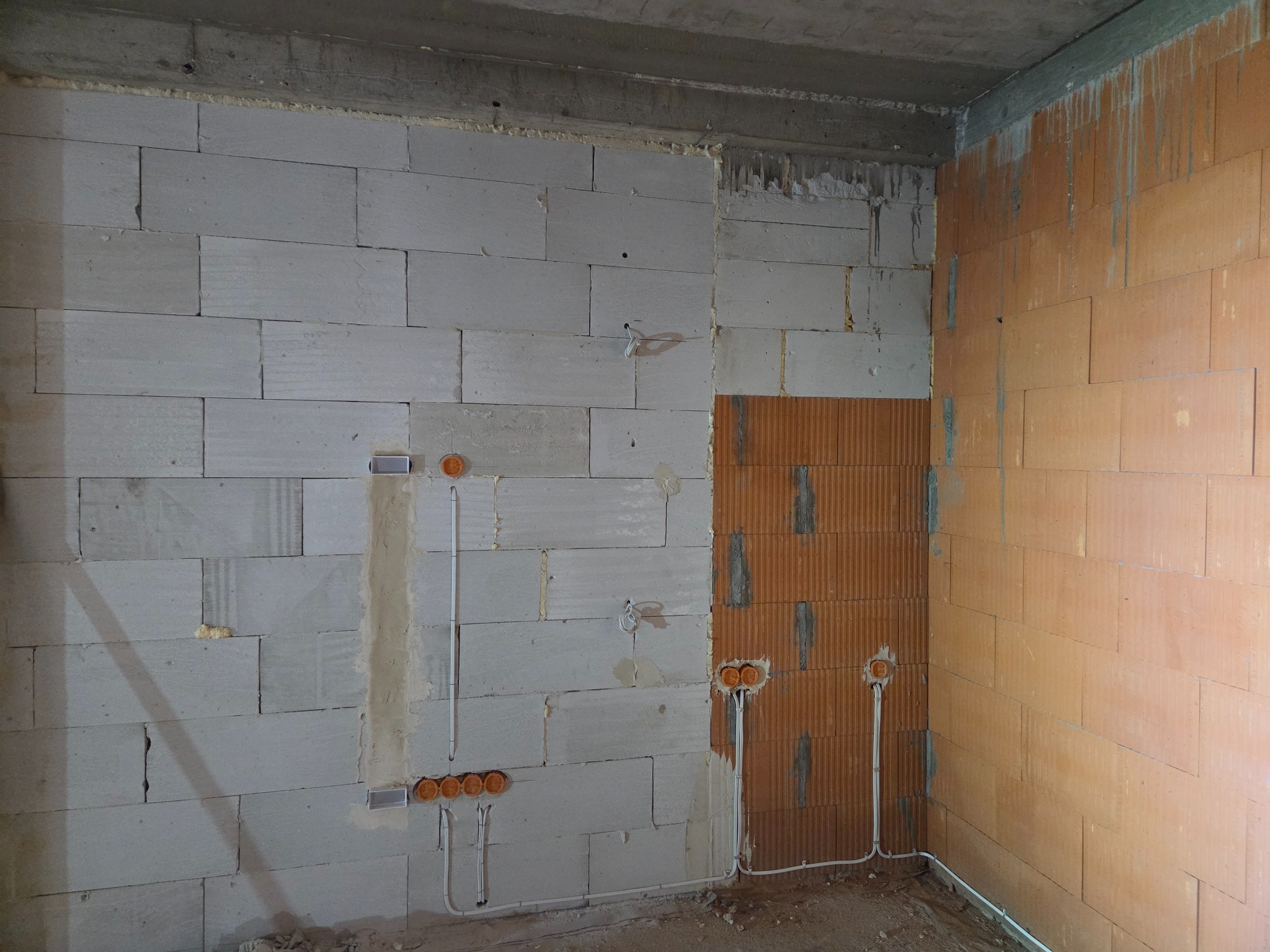 Surowy stan: ściana z pustaków z bruzdami pod instalację elektryczną i puszkami. Widoczne przewody. Fragment ściany z cegły dziurawki. Betonowy strop.