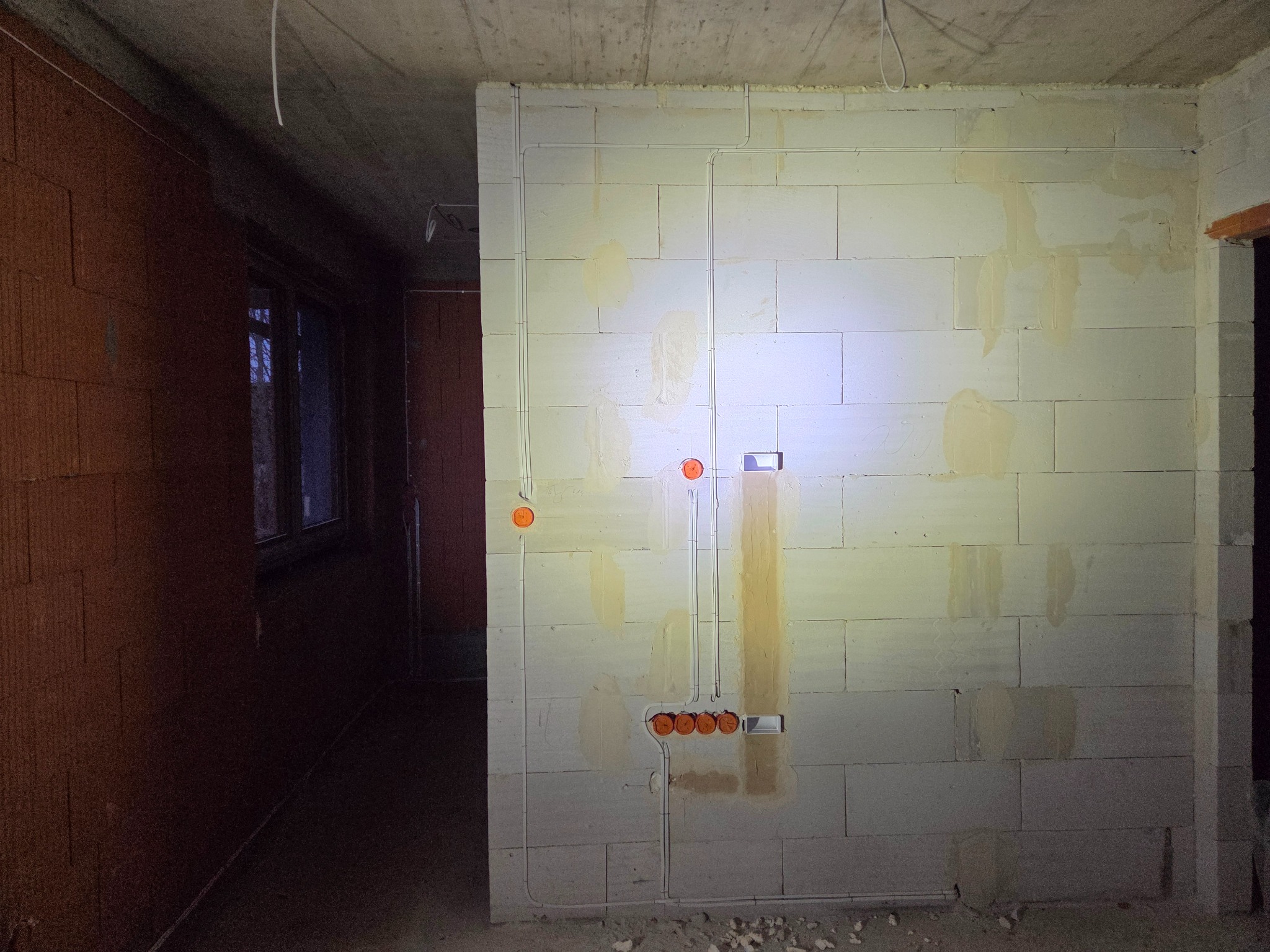 Ściana z betonu komórkowego z rozprowadzoną instalacją elektryczną: kable w peszlach, puszki pod gniazdka i włączniki. Surowy stan, widoczne ślady po zaprawie.