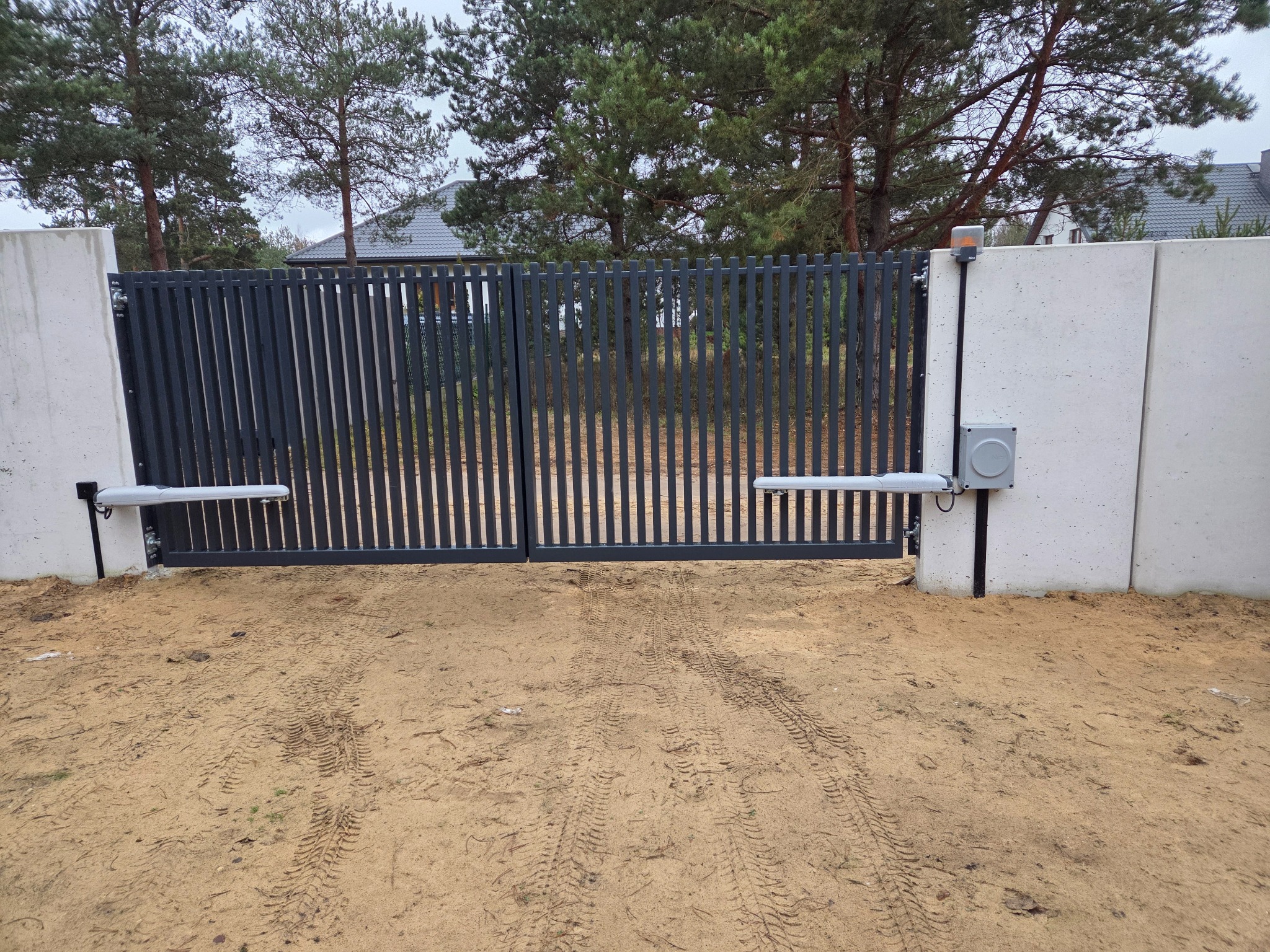 Nowoczesna, automatyczna brama wjazdowa z szarymi, pionowymi panelami, zamontowana na betonowych słupkach. Widoczne siłowniki i lampa ostrzegawcza. Ślady opon na piasku.