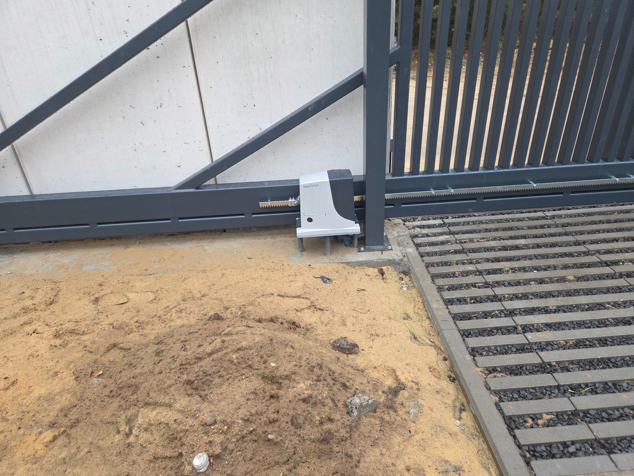 Szara brama przesuwna z zamontowanym napędem automatycznym Nice Robus, widoczna listwa zębata i betonowe podłoże.