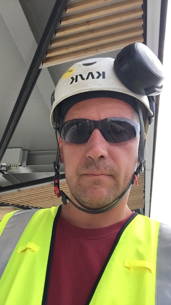 Selfie hydraulika w okularach przeciwsłonecznych, kasku z nausznikami i kamizelce odblaskowej na tle instalacji pod sufitem.