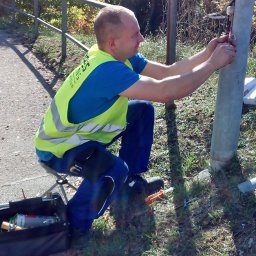 D-instal.NET Bartosz - Elektryk w kamizelce montuje oświetlenie uliczne przy drodze. Widoczna skrzynka z narzędziami i fragment chodnika. Słoneczny dzień.