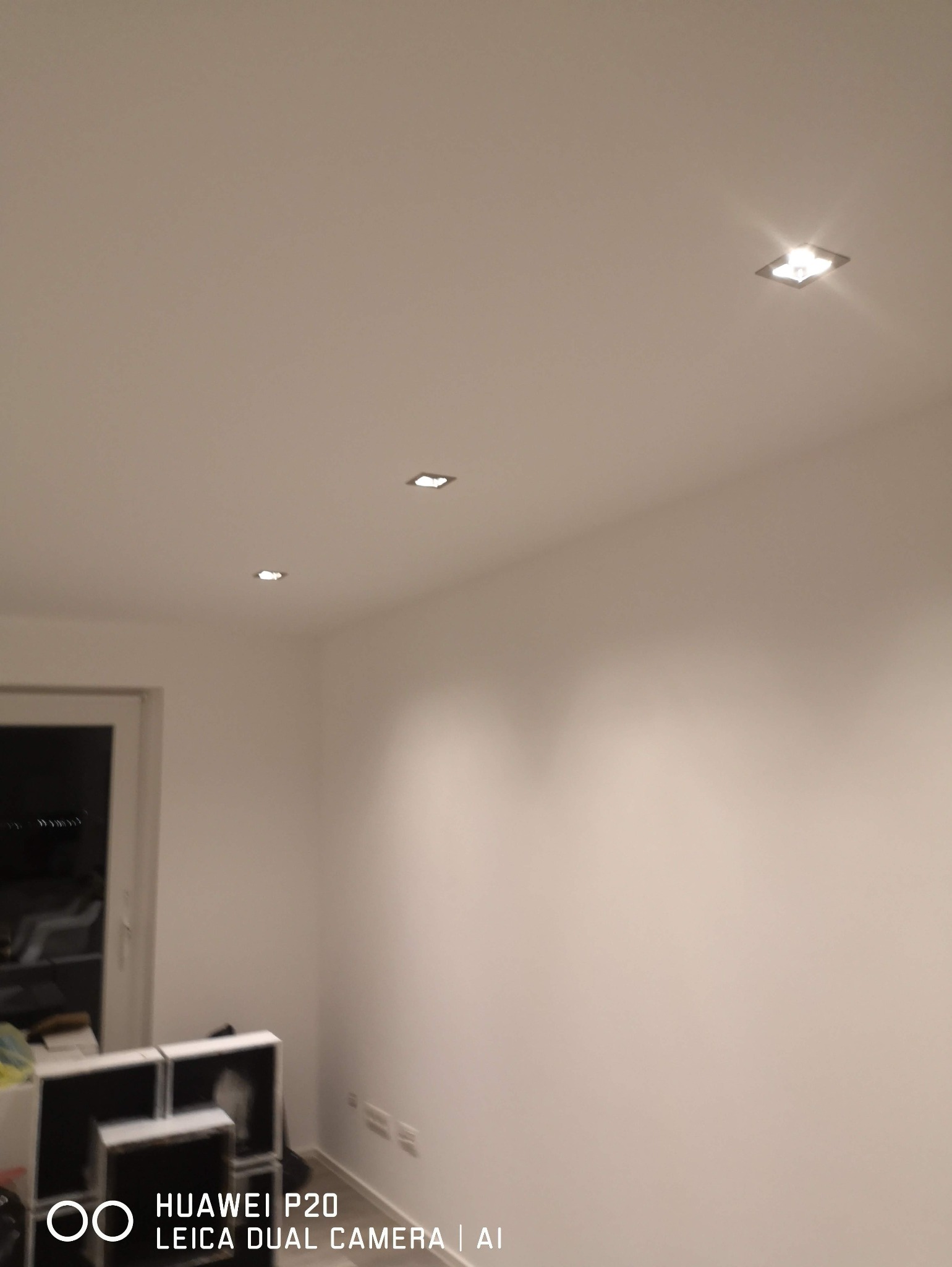 Białe wnętrze z trzema kwadratowymi lampami sufitowymi LED, świecącymi ciepłym światłem. Widoczny fragment drzwi i niezidentyfikowane obiekty w rogu.