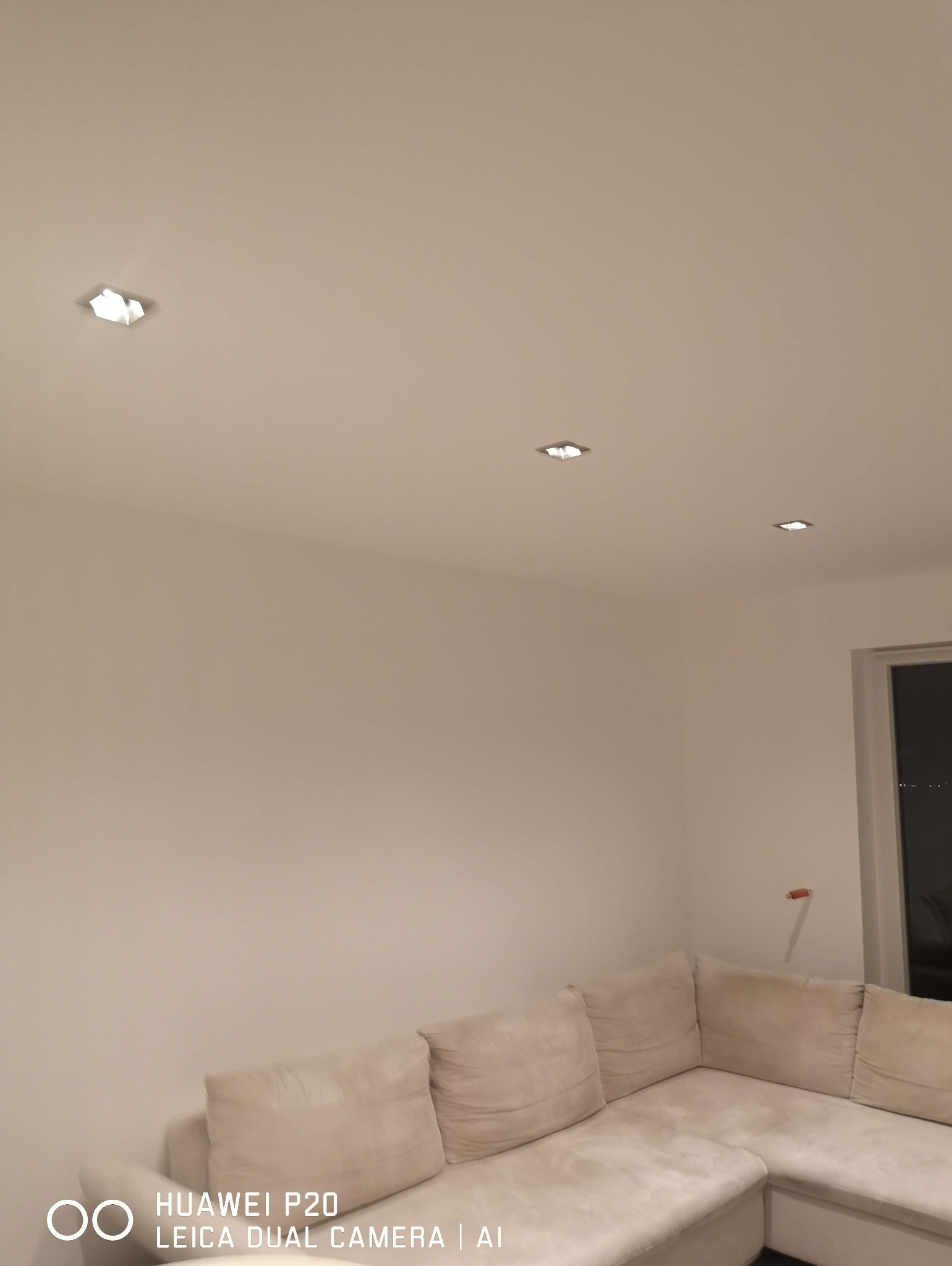 Jasne wnętrze z trzema punktowymi lampami sufitowymi, oświetlającymi kremową sofę narożną. Minimalistyczny design, ciepłe światło.