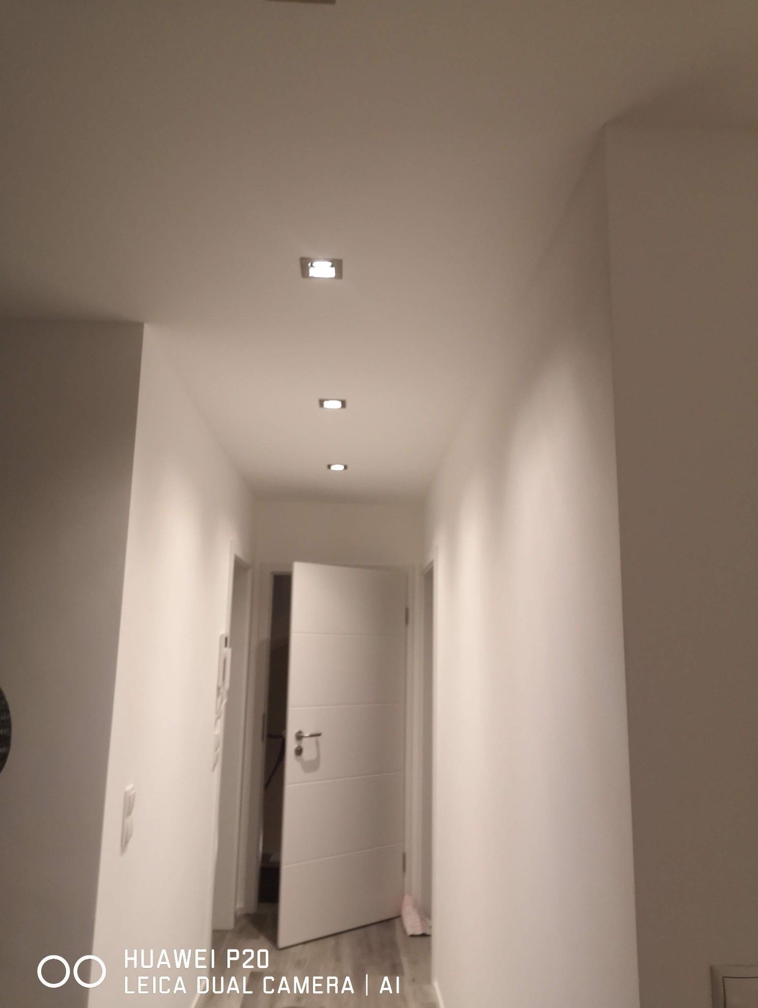 Korytarz z trzema kwadratowymi lampami sufitowymi LED, białe ściany, otwarte drzwi na końcu korytarza. Minimalistyczny design oświetlenia.
