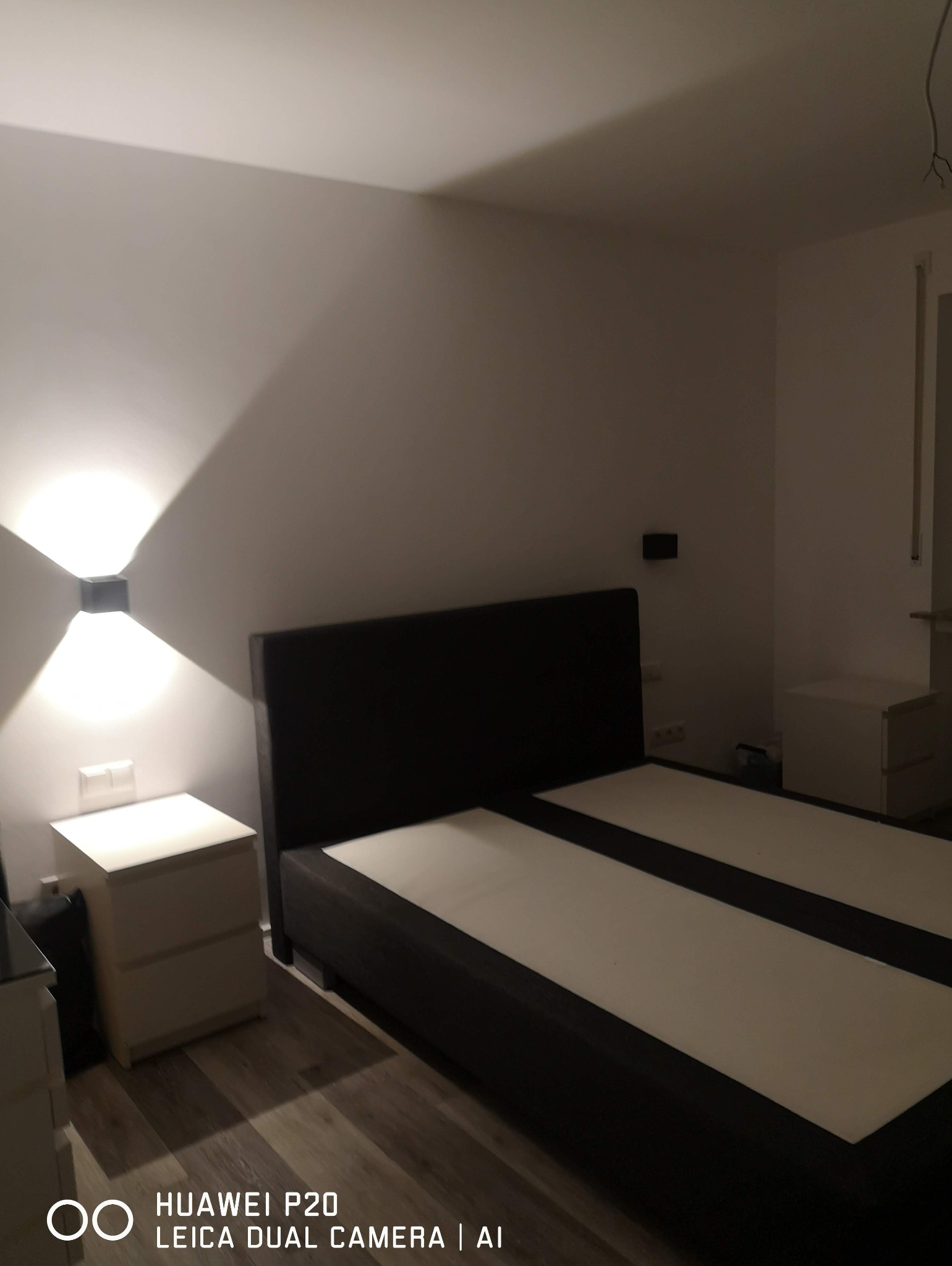 Nowoczesna sypialnia z podwójnym łóżkiem i zamontowanym na ścianie, kwadratowym oświetleniem LED, rzucającym symetryczne światło na ścianę. Widoczny kontakt i szafka nocna.