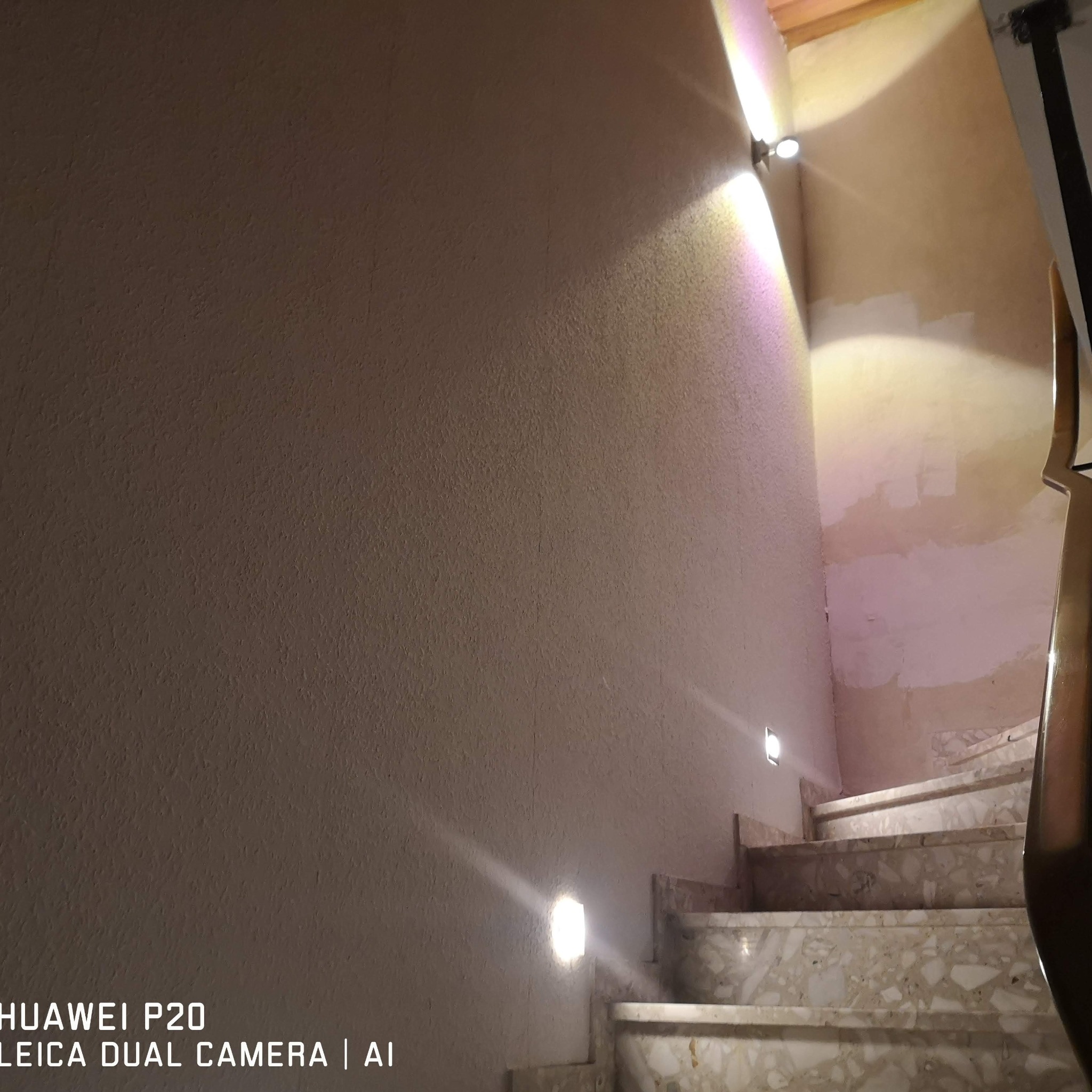 Klatka schodowa z marmurowymi schodami oświetlona przez dwa nowoczesne, ścienne punkty świetlne, rzucające snopy światła na teksturowaną ścianę.