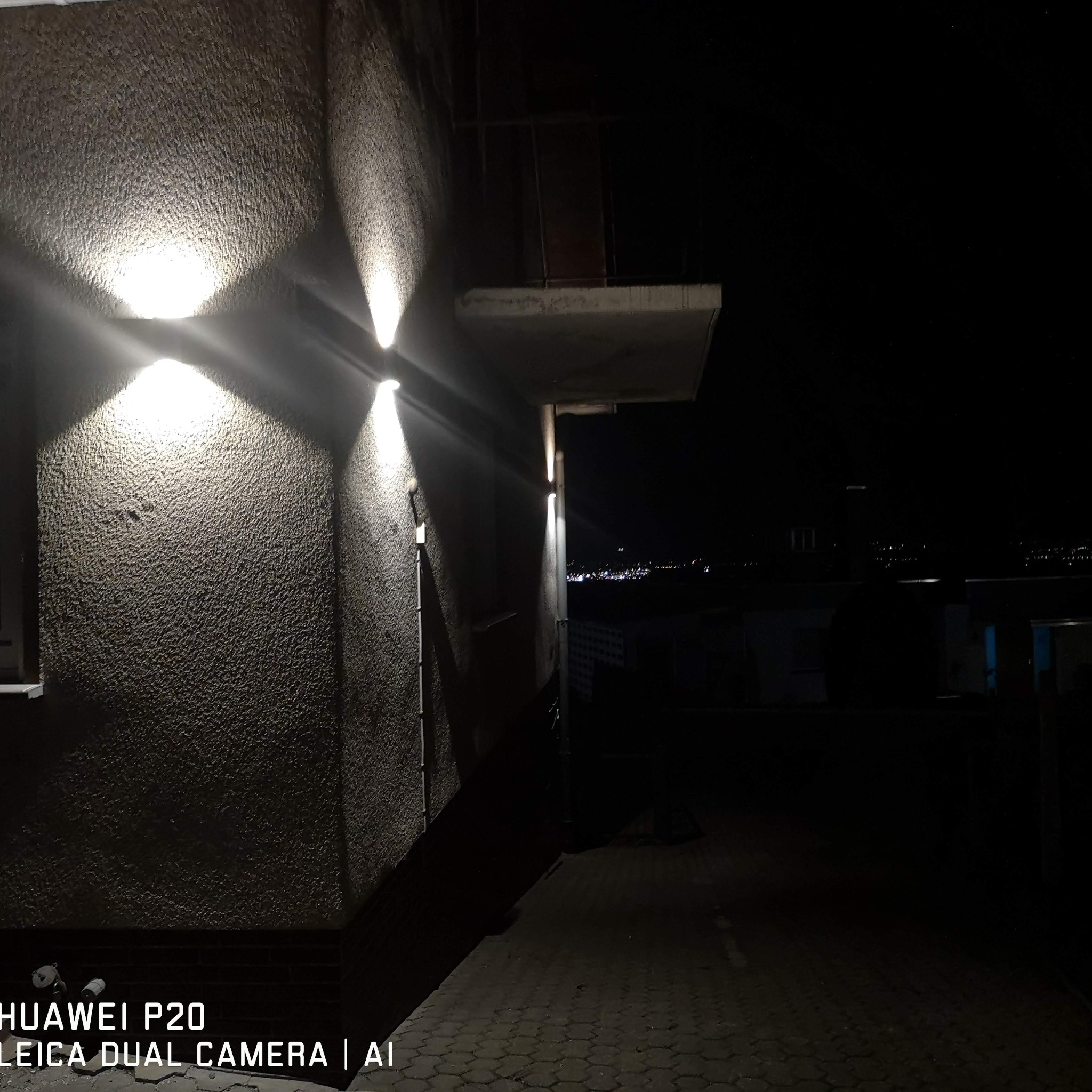 Dwa reflektory elewacyjne oświetlają szorstką ścianę budynku nocą. W tle widoczne światła miasta.