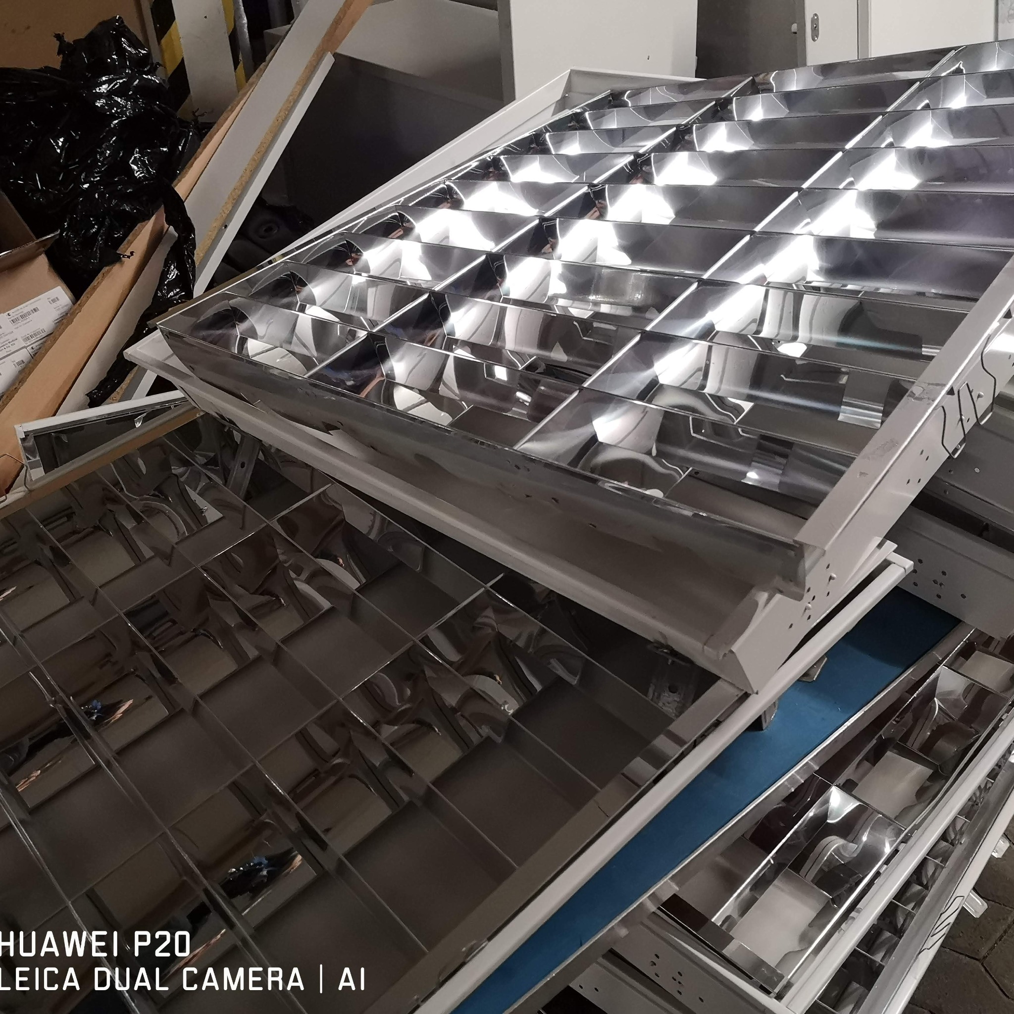 Stos srebrnych, metalowych opraw oświetleniowych z rastrem, ułożonych jedna na drugiej. Widoczne elementy montażowe i odbicia światła.