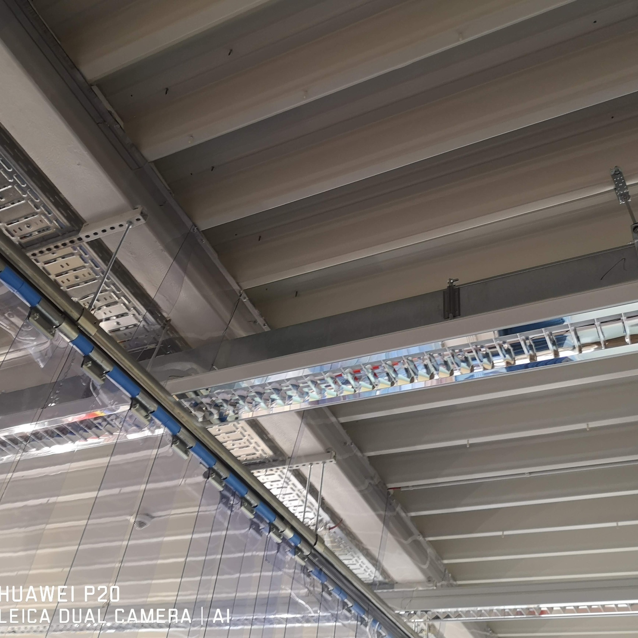 Sufit przemysłowy z widocznymi instalacjami: koryta kablowe, rury z niebieskimi oznaczeniami i oświetlenie rastrowe. Ujęcie z dołu, perspektywa zbieżna.