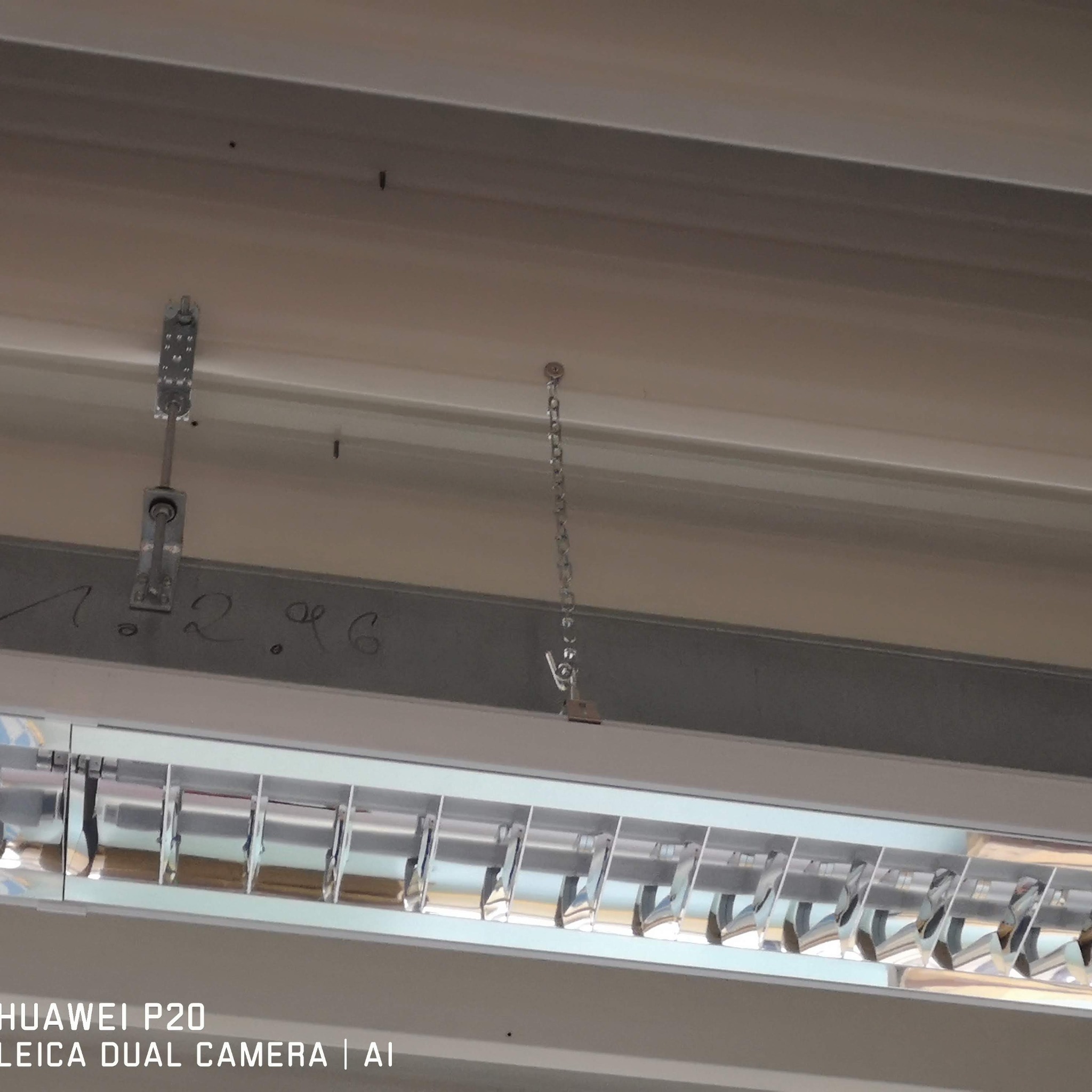 Zamontowana, podwieszana lampa rastrowa z widocznymi elementami mocującymi do sufitu, w tym łańcuszkiem. Na belce widoczny napis '1.2.96'.