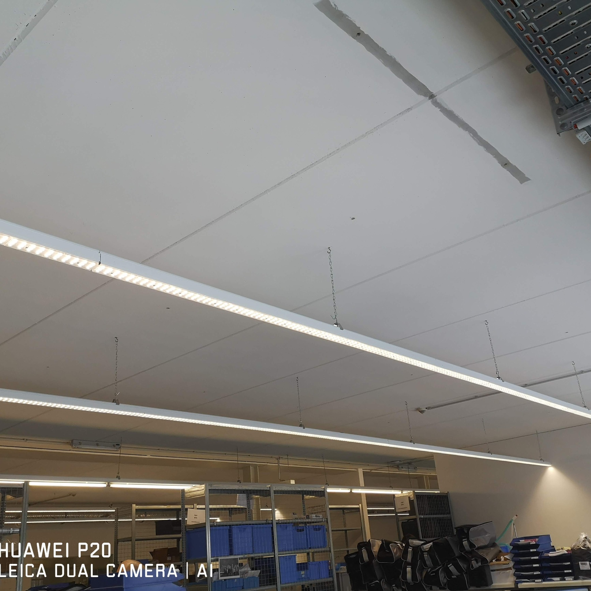 Długie, wiszące lampy LED oświetlają magazyn z regałami i niebieskimi pojemnikami. Widoczne łączenia płyt sufitowych i łańcuchy podtrzymujące oświetlenie.