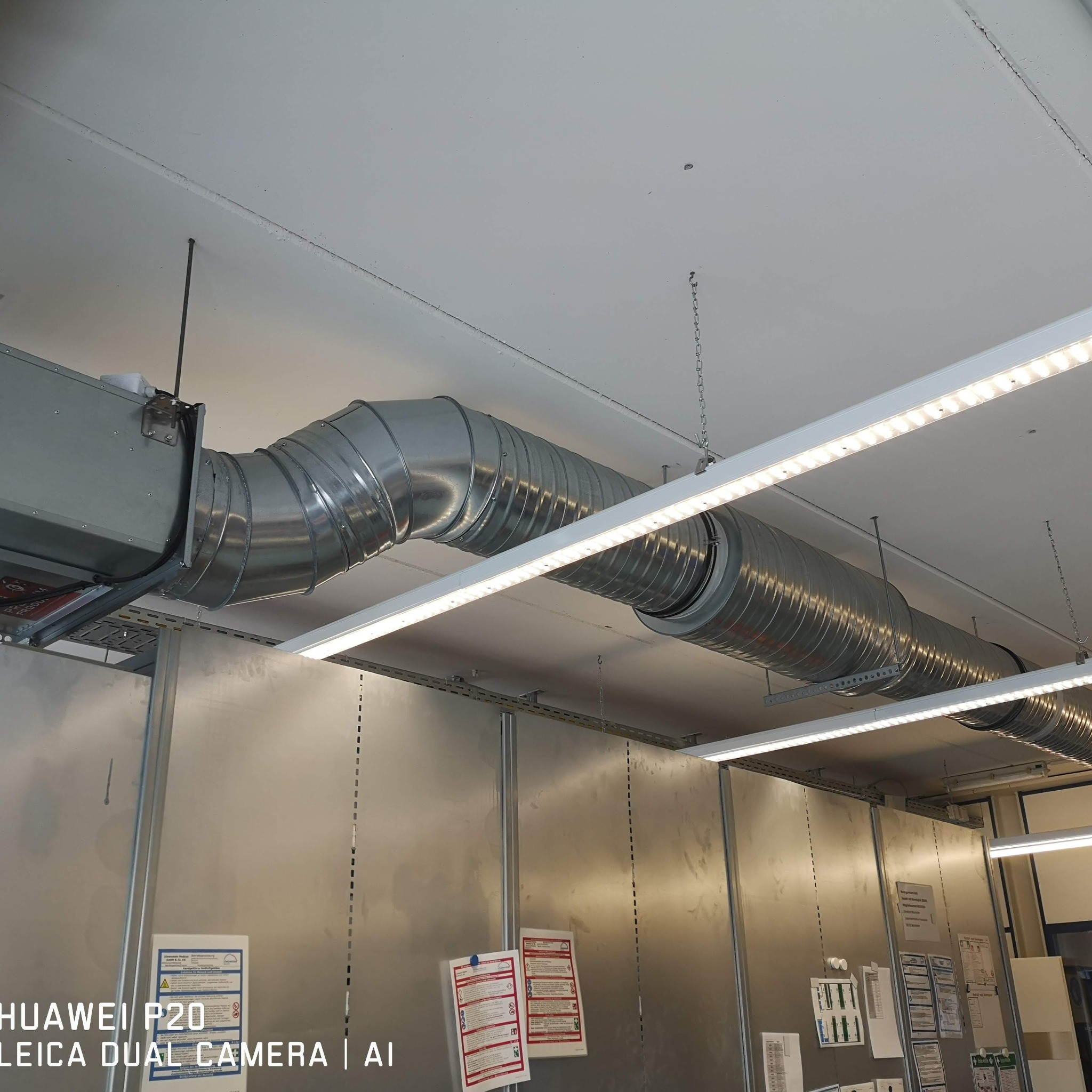 Zamontowany system wentylacyjny z metalowych rur pod sufitem, oświetlenie LED w tle. Widoczne elementy mocujące i tablice informacyjne na ścianie.