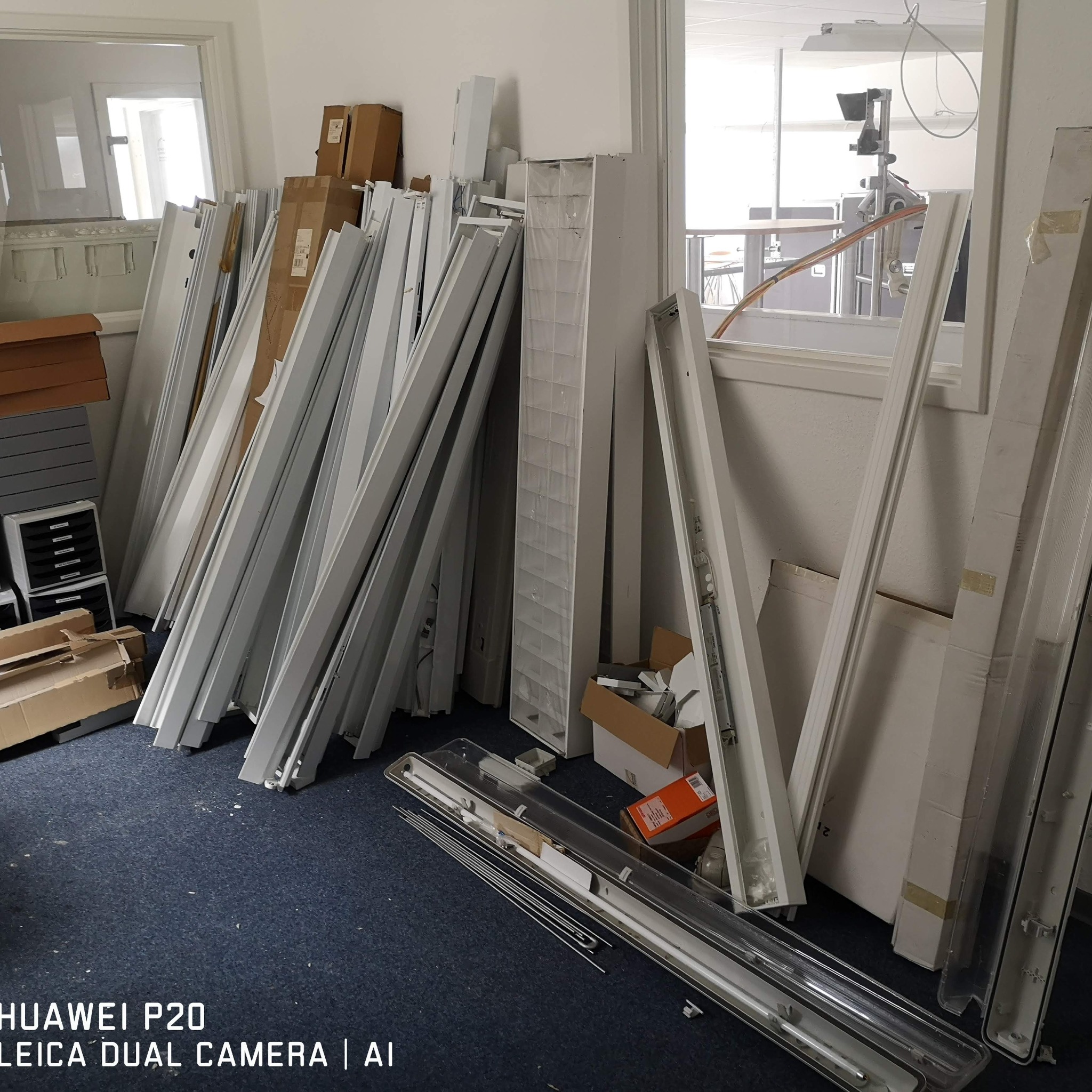 Składowisko elementów oświetleniowych: białe obudowy lamp, profile i opakowania kartonowe, tworzące nieuporządkowany stos w pomieszczeniu z oknem.