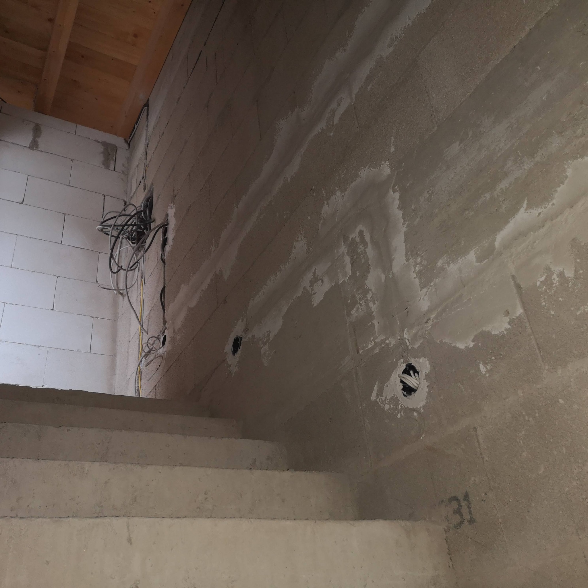 Surowa ściana z kablami wychodzącymi z otworu, widoczne ślady po szpachlowaniu. W tle fragment schodów i drewniany strop.