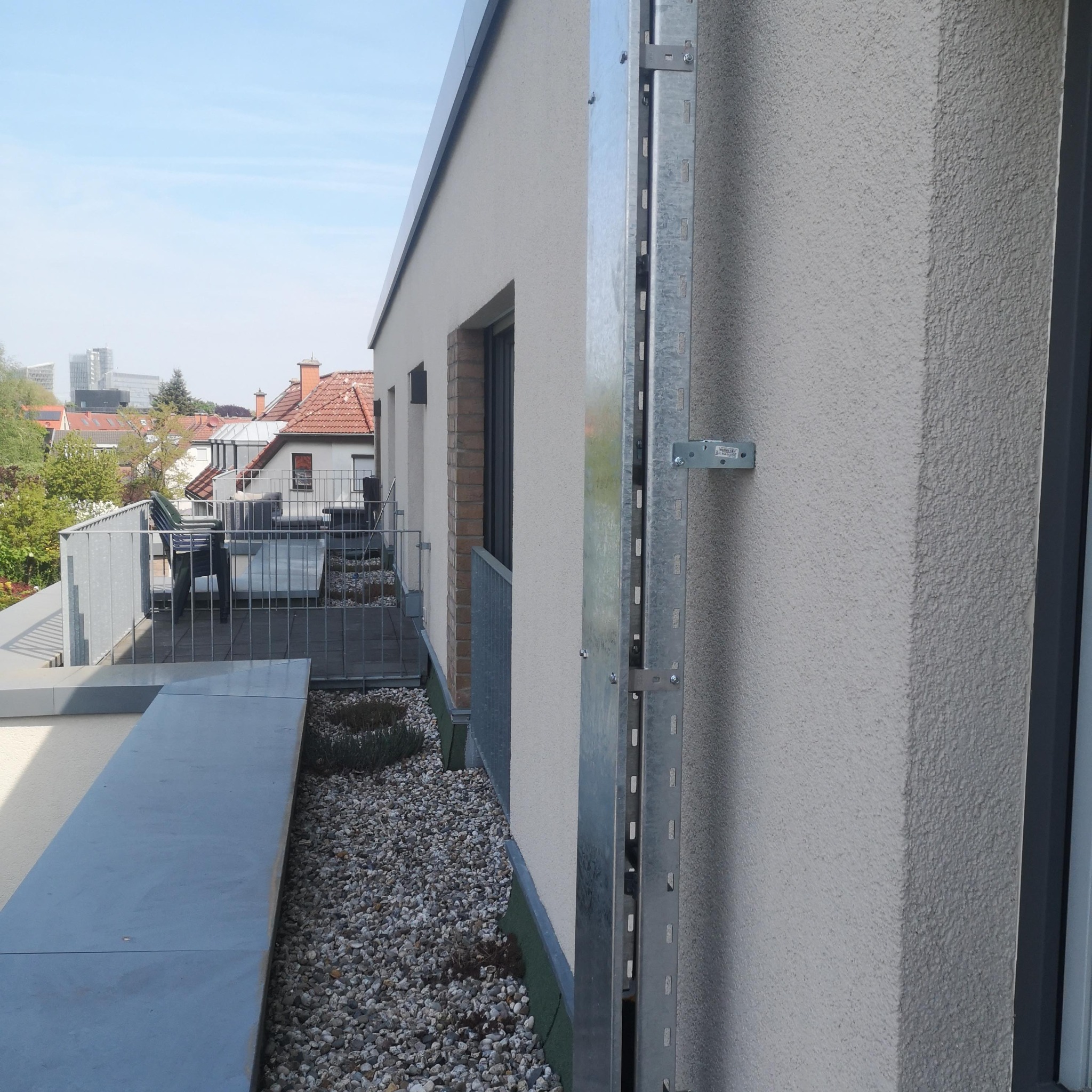 Metalowa listwa montażowa przymocowana do elewacji budynku, widok z zewnątrz, z balkonem w tle i elementami instalacji na dachu.