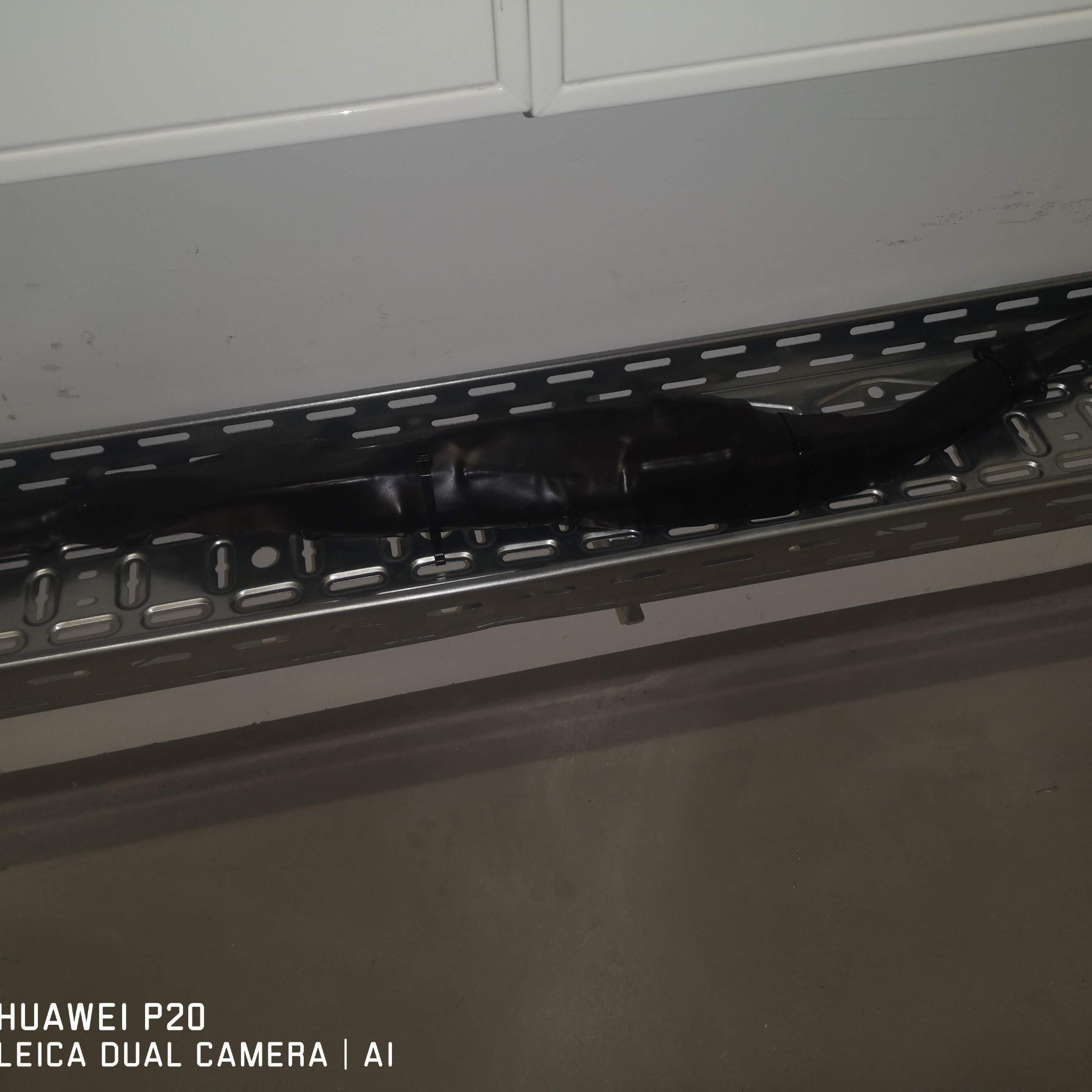 Kable elektryczne zabezpieczone czarną taśmą izolacyjną, ułożone w metalowej korycie kablowej, zamontowanej pod sufitem. Widoczne otwory wentylacyjne w korycie.