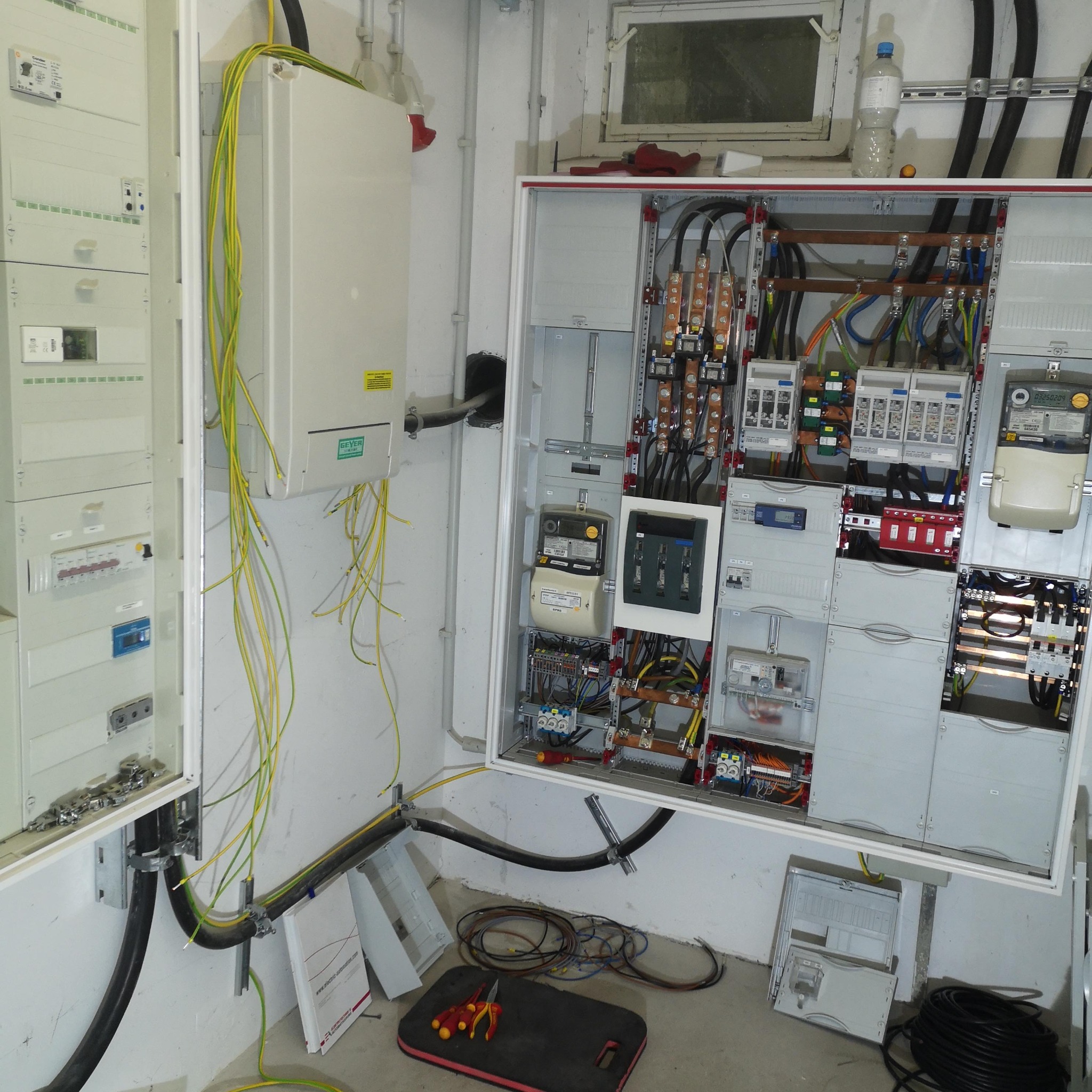 Rozdzielnia elektryczna z otwartą obudową, widoczne kable, bezpieczniki i liczniki. Na podłodze narzędzia i fragmenty obudowy. Ściany białe, surowe wykończenie.
