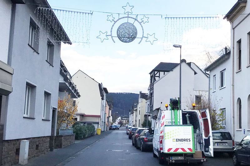 Ulica w miasteczku z dekoracjami świątecznymi zawieszonymi nad drogą. Na drugim planie góry. Samochód z podnośnikiem i logo firmy oświetleniowej.