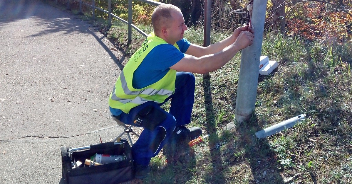 Elektryk w kamizelce montuje oświetlenie uliczne przy drodze. Widoczna skrzynka z narzędziami i fragment chodnika. Słoneczny dzień.