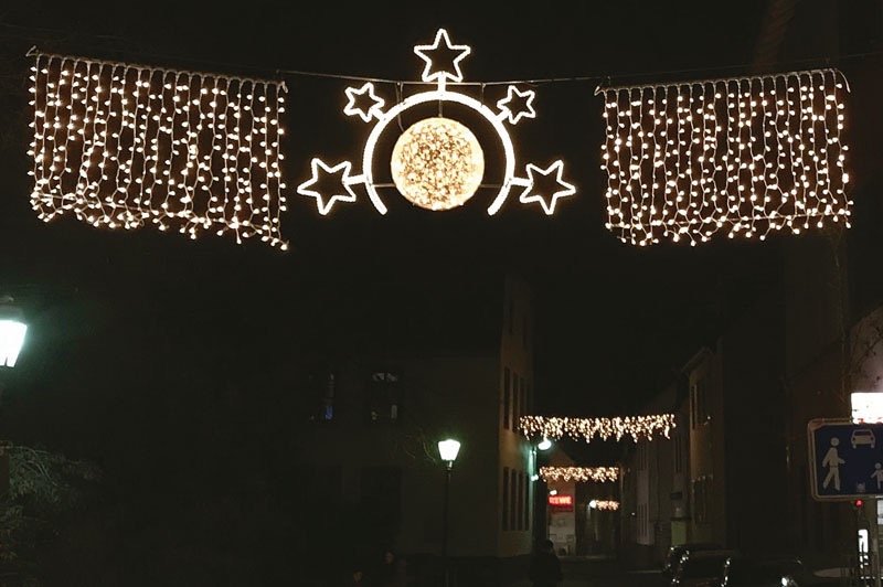 Świąteczna dekoracja uliczna: girlandy z lampek w kształcie gwiazd i kuli, oświetlające ciemną ulicę z budynkami w tle. Nocna sceneria, ciepłe światło.