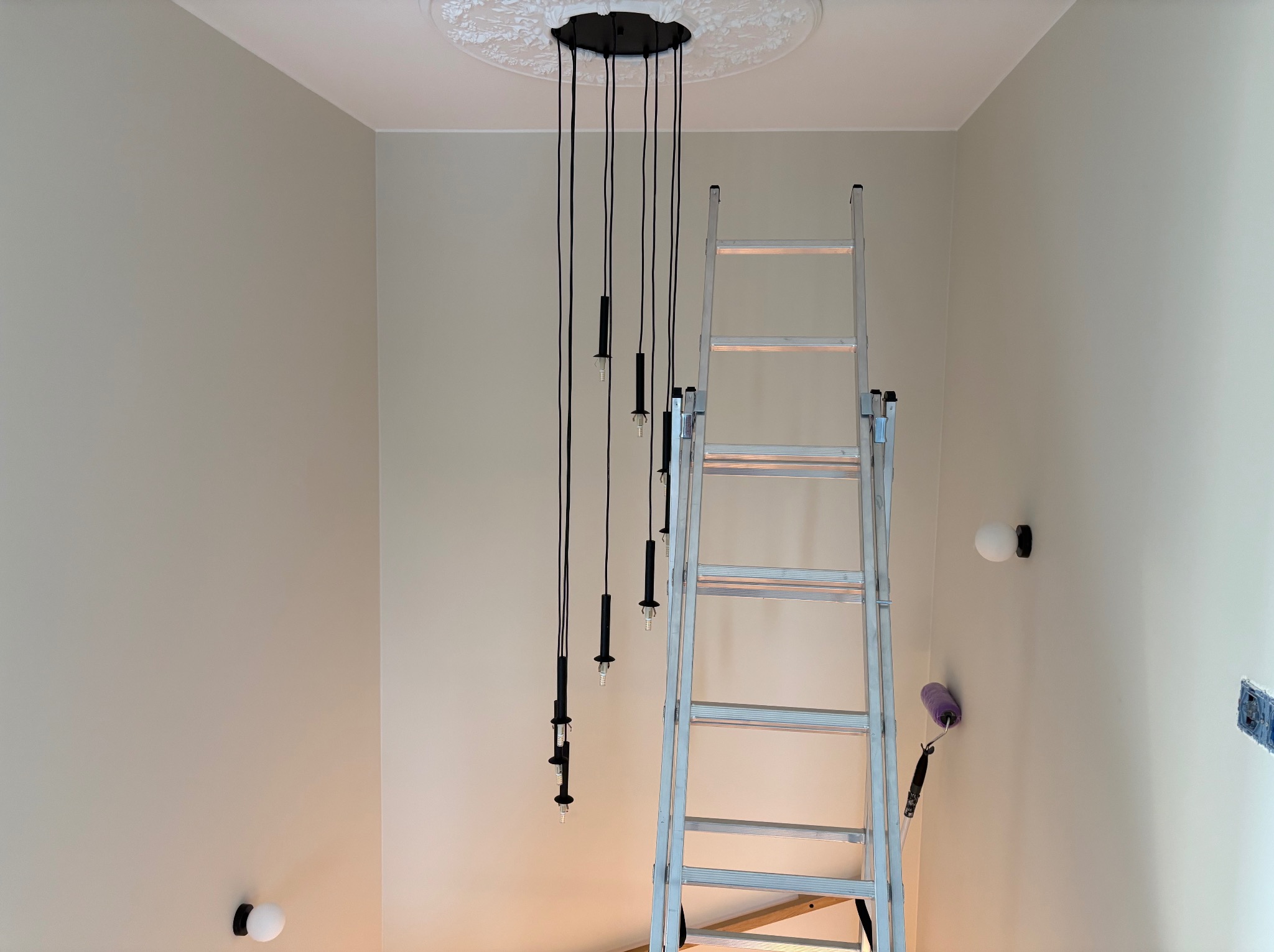 Montaż lamp wiszących z czarnymi kablami i dekoracyjną rozetą sufitową. Aluminiowa drabina oparta o ścianę. Widoczne kable i puszka elektryczna.