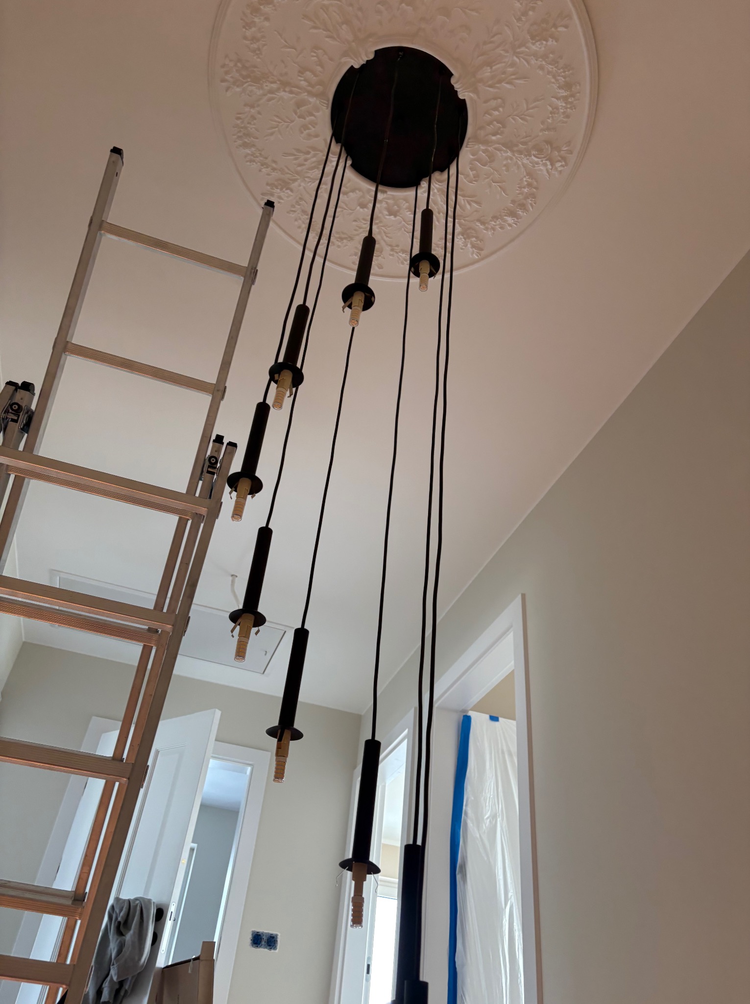 Montaż czarnej, wiszącej lampy z wieloma źródłami światła na tle białego sufitu z dekoracyjną rozetą. Widoczna drabina i przygotowane pomieszczenie.