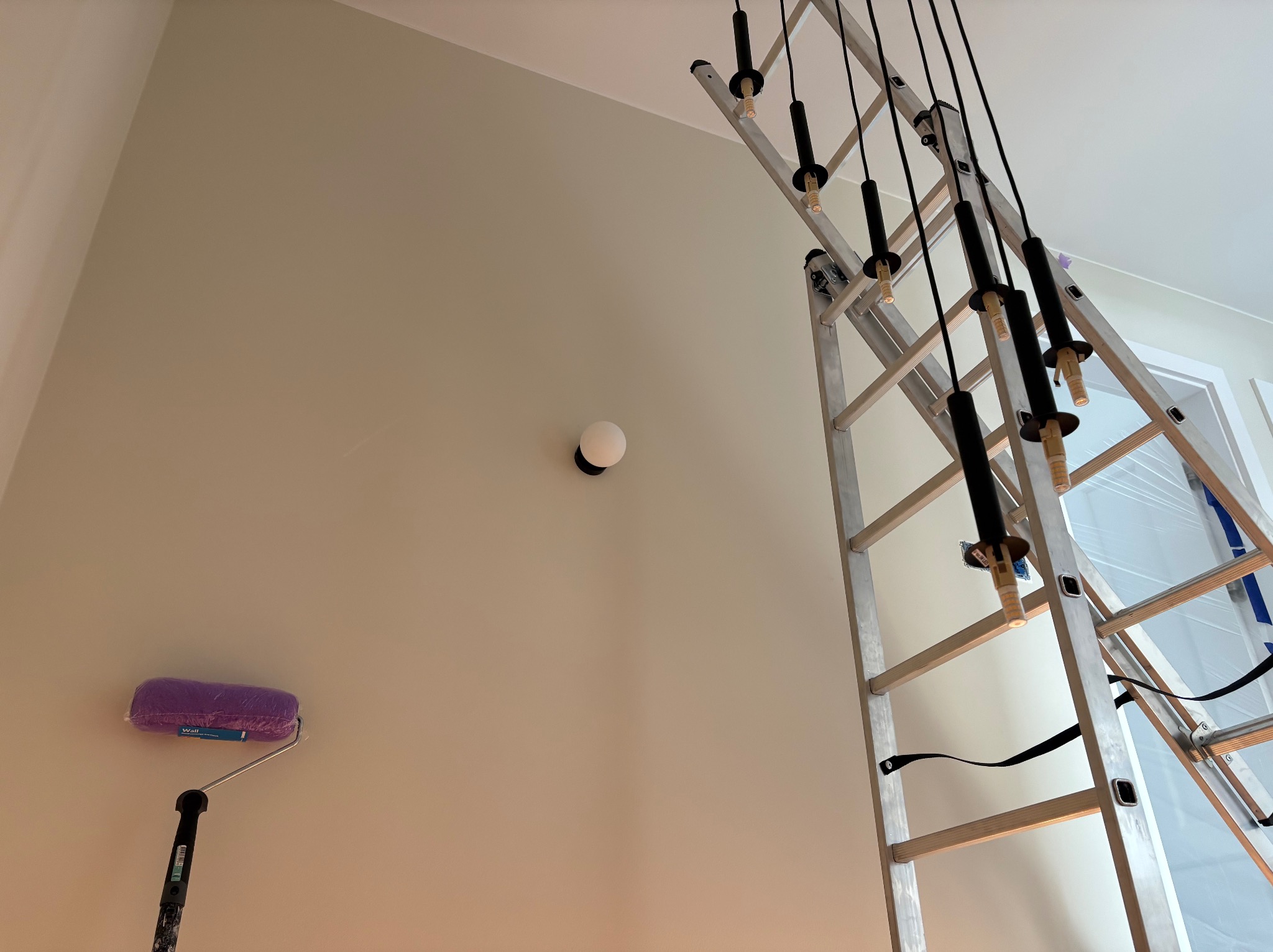 Malowanie ścian w jasnym odcieniu. Widoczna drabina z zamontowanym oświetleniem i wałek do malowania. Świeżo pomalowana ściana z zamontowanym kinkietem.
