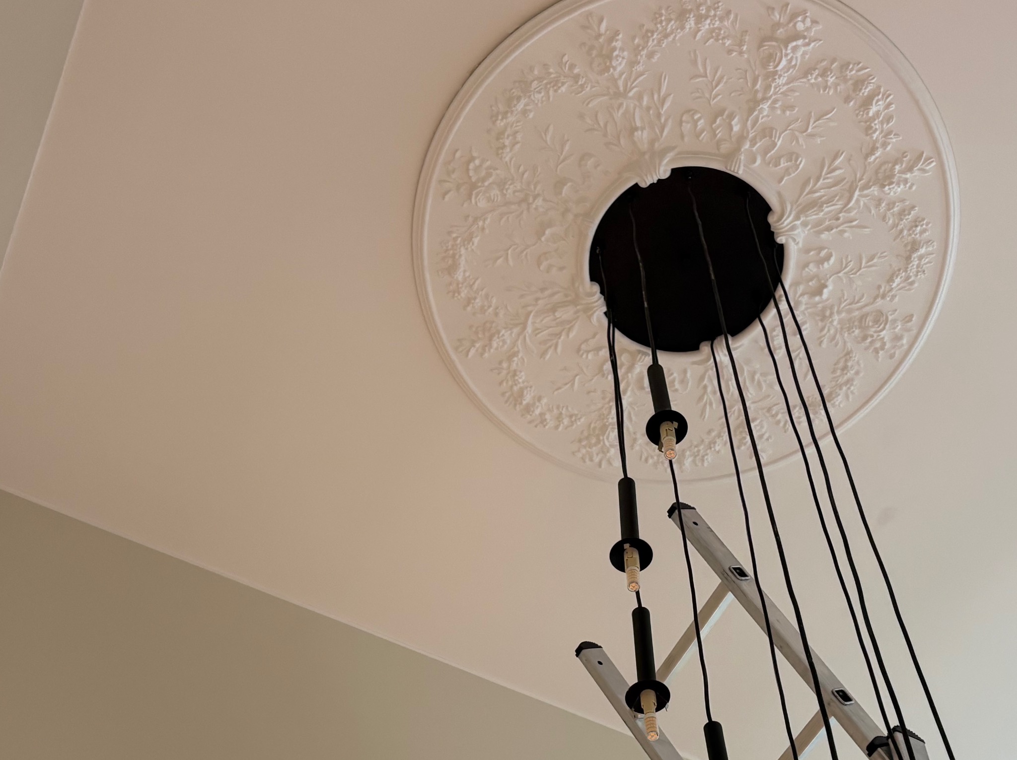 Dekoracyjny plafon z rozetą i zwisającymi przewodami oświetleniowymi, częściowo zasłonięty przez rozłożoną drabinę aluminiową. Widok z dołu.